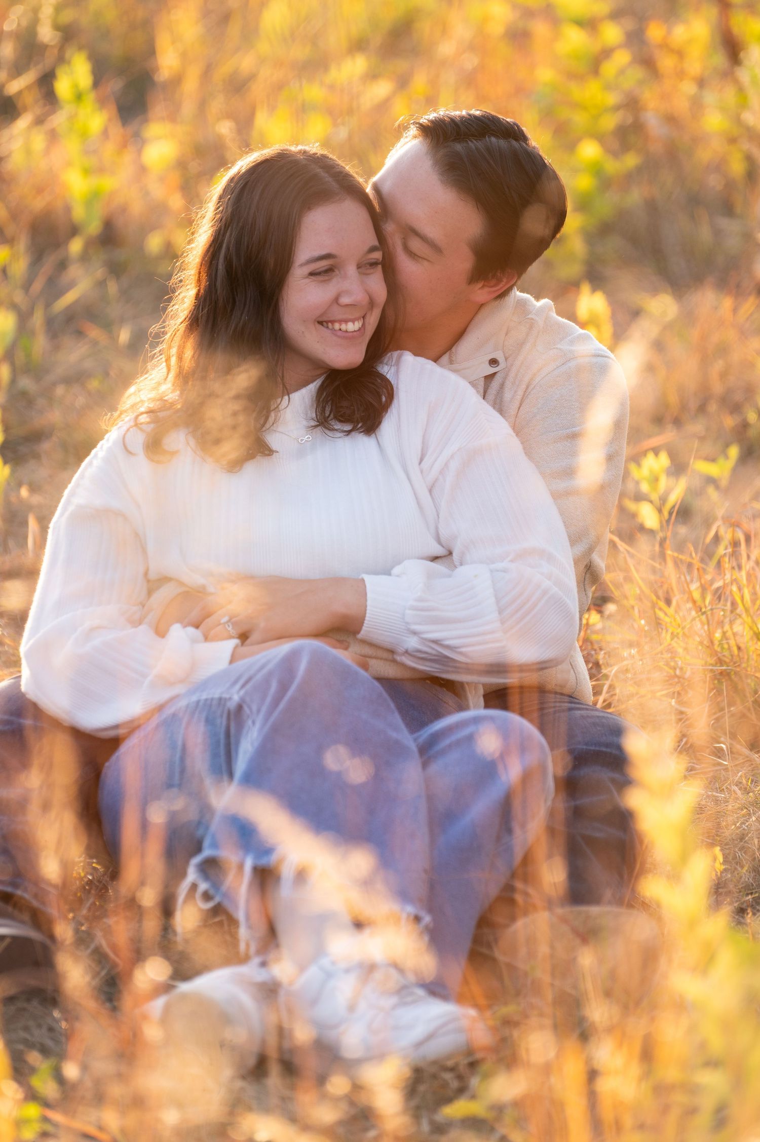 A romantic couple embraces while sitting in a sun-lit field of golden grass during sunset.