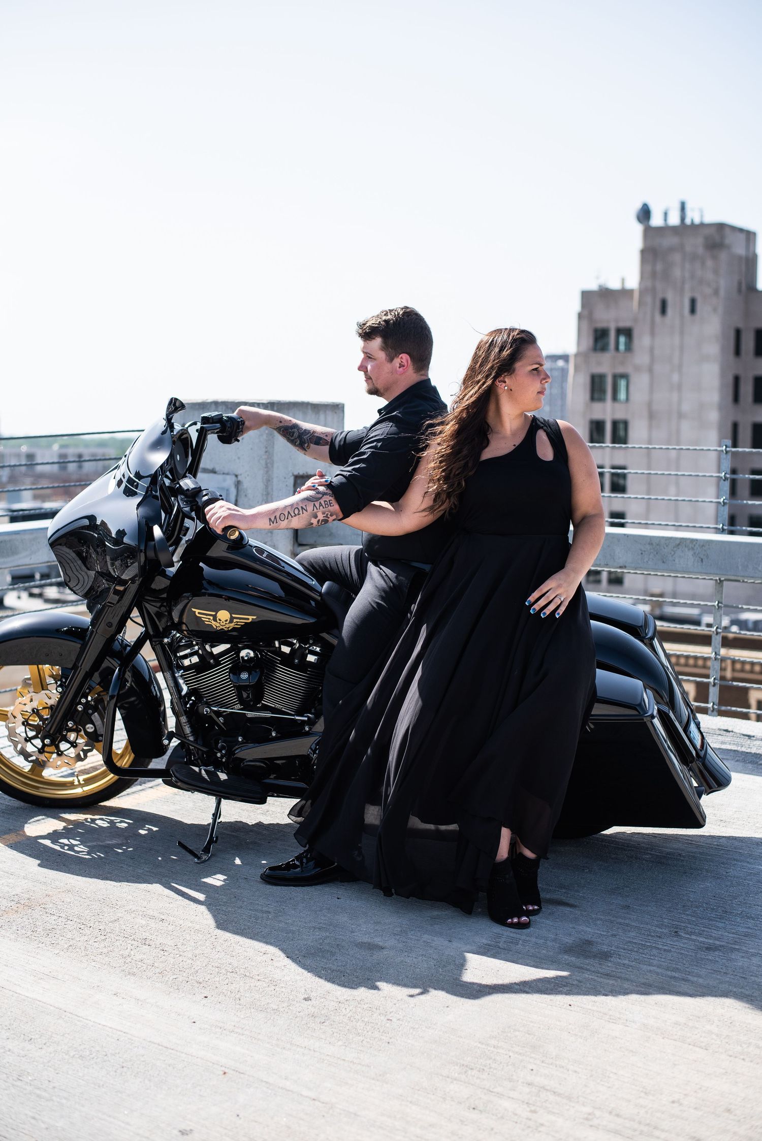 Couple poses with a black Harley Davidson motorcycle on an urban rooftop.