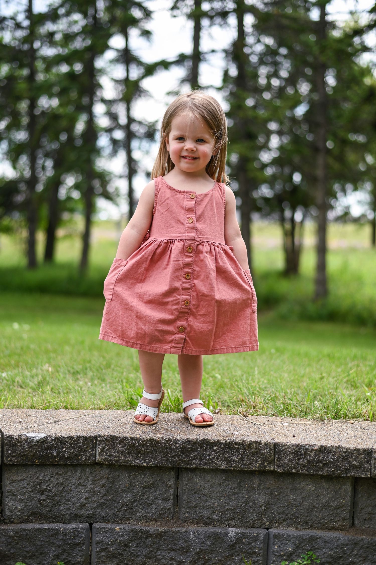 Young girl in pink dress sitting on concrete ledge with trees in background.