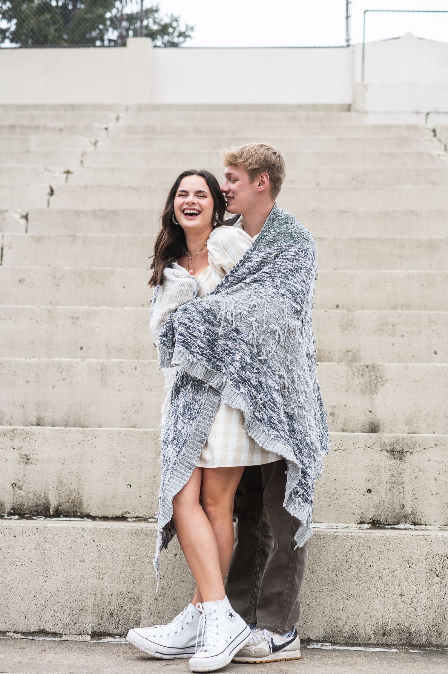 A couple wrapped in a gray blanket shares a laugh on concrete steps.
