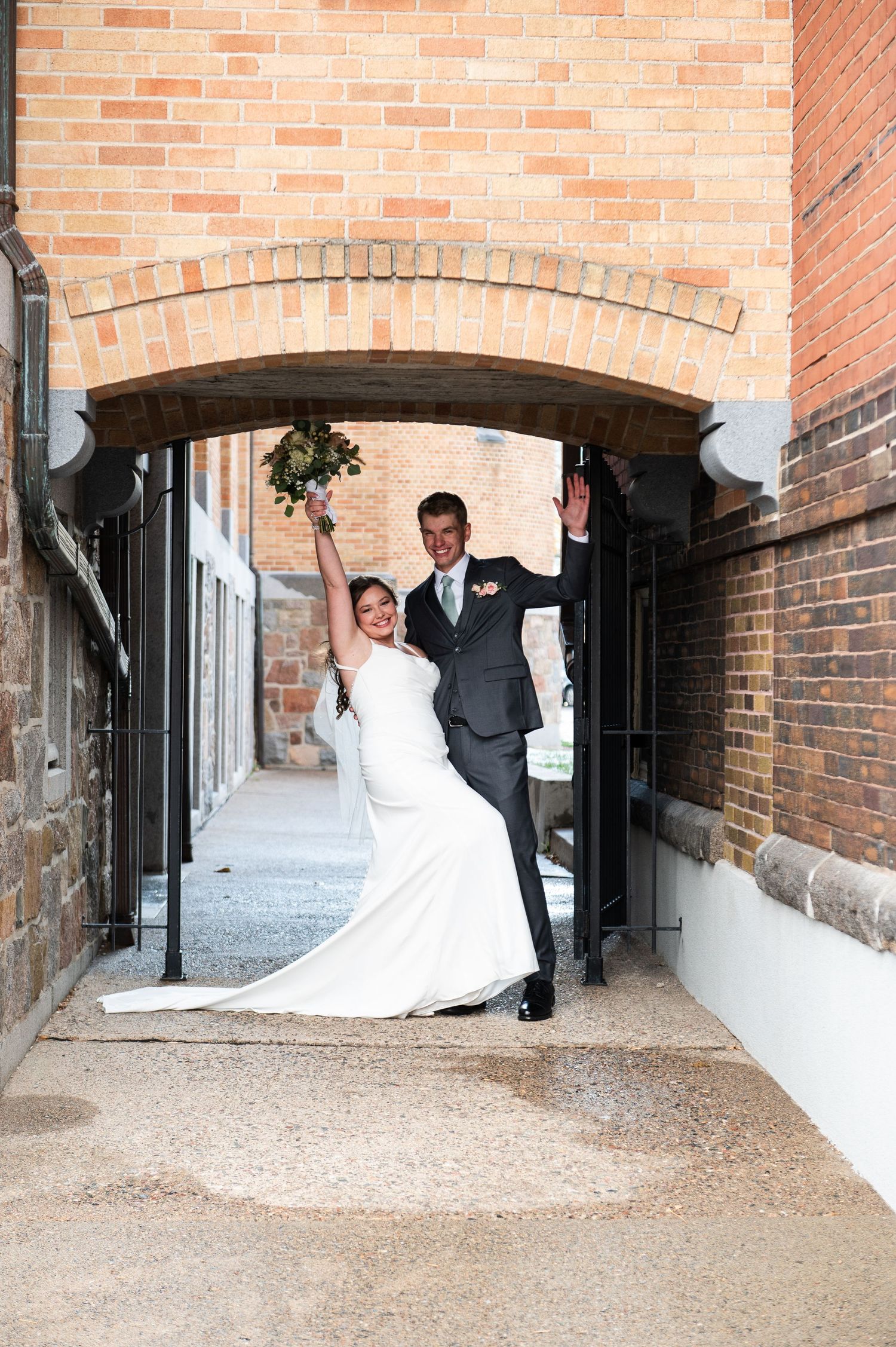 A couple poses under a brick archway in formal wedding attire.