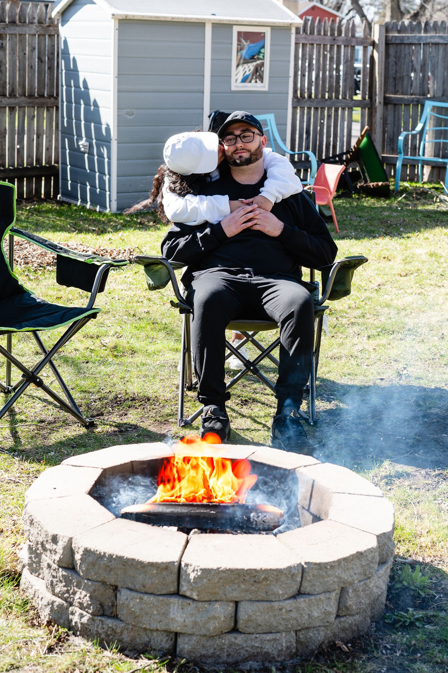 People sit in camping chairs around a stone fire pit with bright flames in a backyard setting.