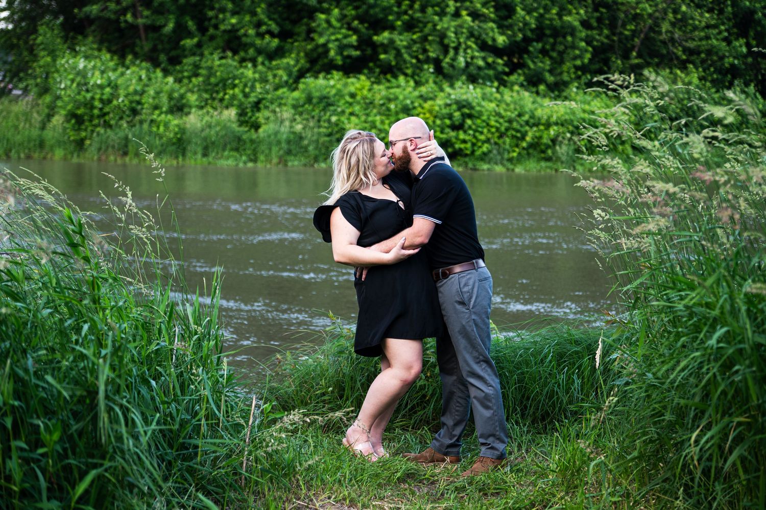 A couple in dark clothing embraces next to a flowing river surrounded by green vegetation during summer.