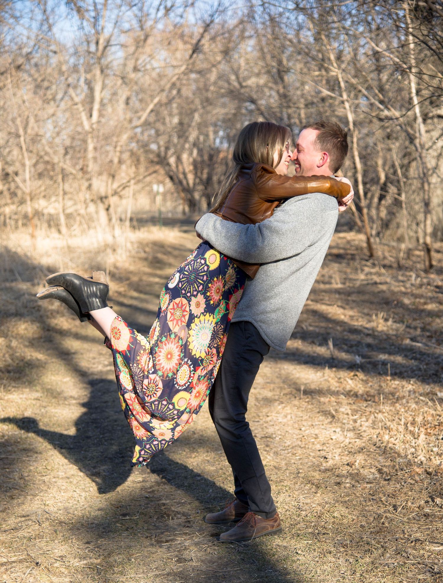 A joyful embrace between two people on a wooded trail, with the person in a floral dress being lifted into the air.
