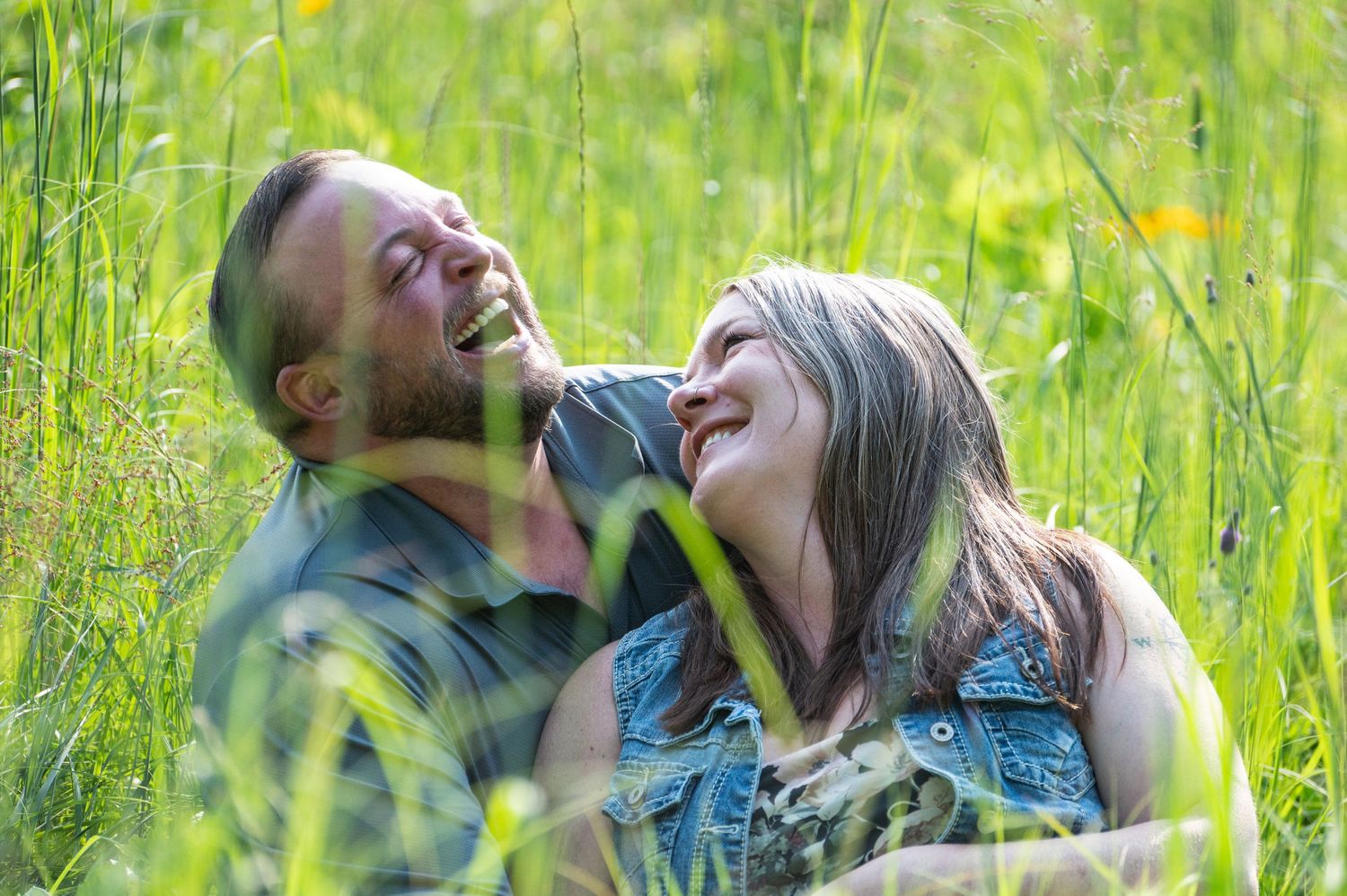 A couple laughs together while lying in tall green grass on a sunny day.