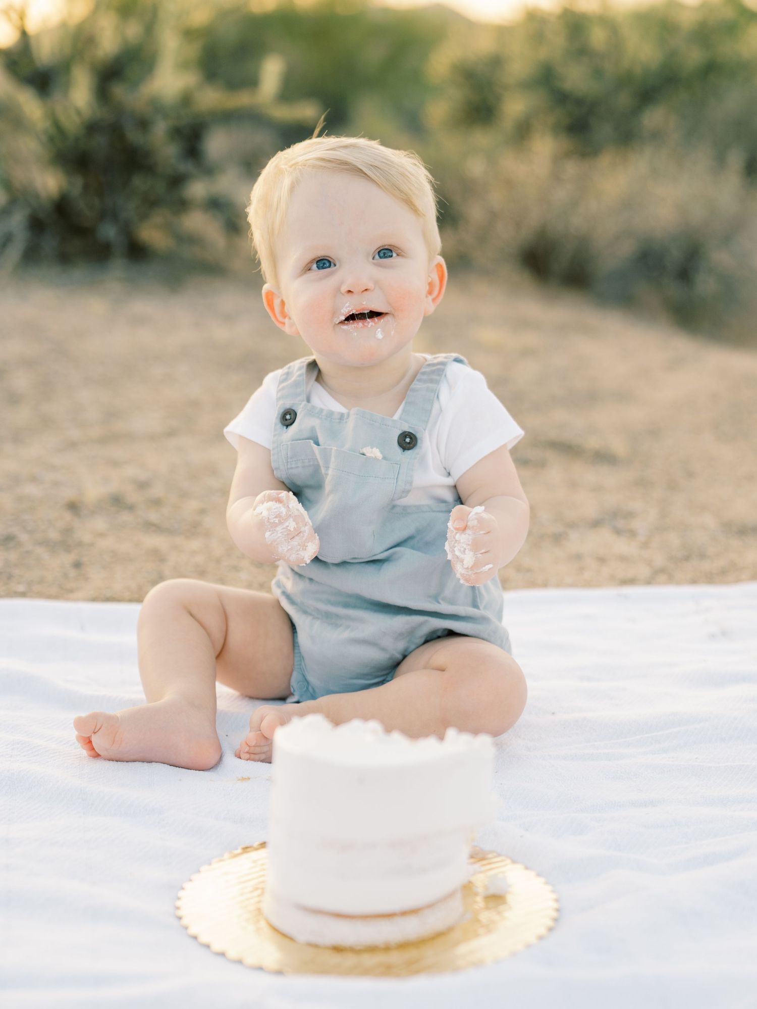 Joyful baby boy in blue overalls celebrating first birthday smash cake outdoors at sunset.