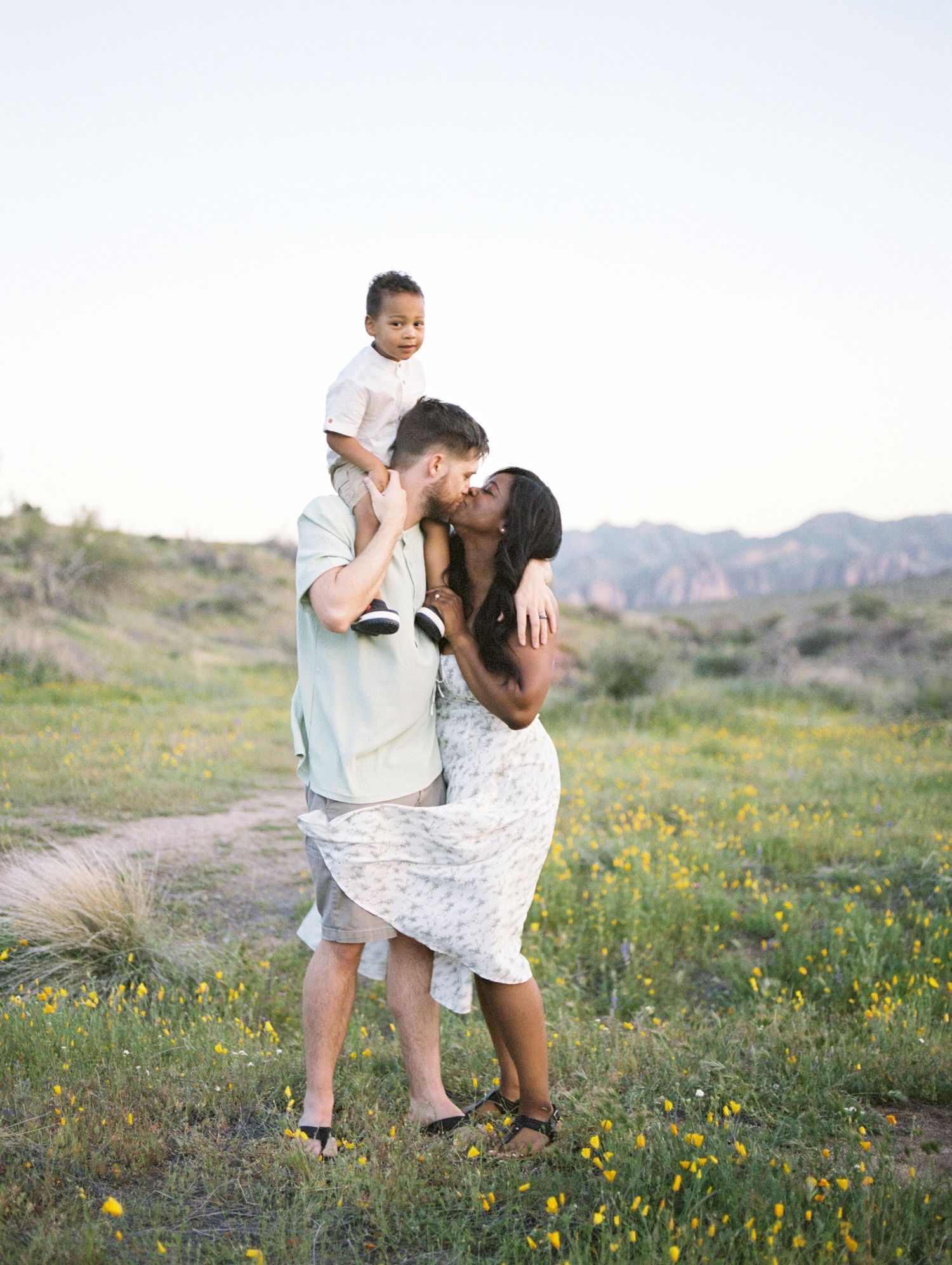 A couple kisses romantically in a wildflower meadow while their toddler sits on dad's shoulders.