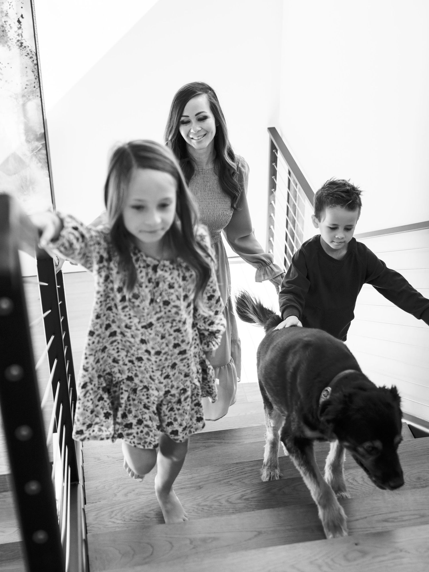 Black and white photo of mother, two children, and dog walking down modern staircase together.