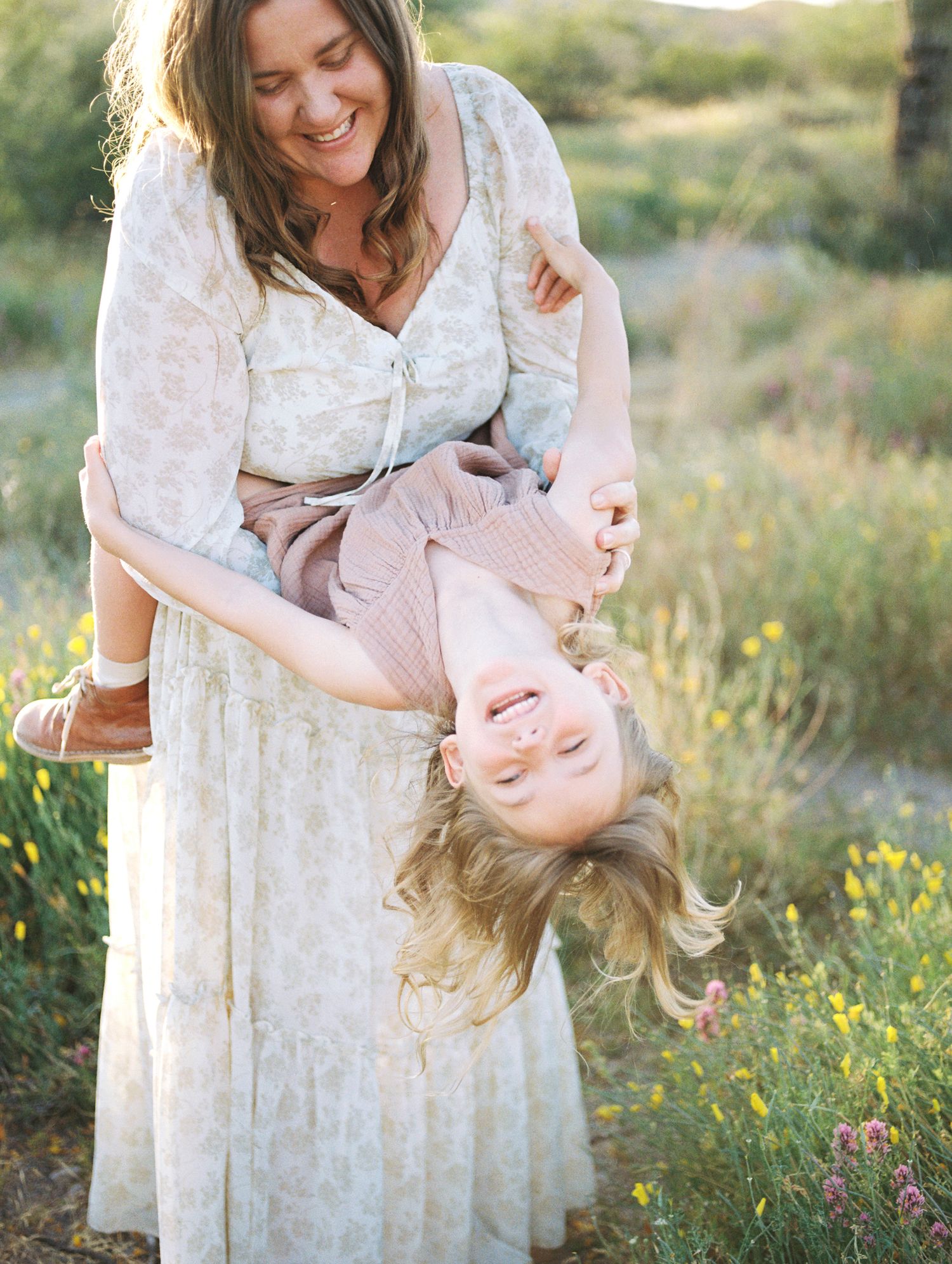 Laughing mother playfully holds young daughter upside down in wildflower meadow.