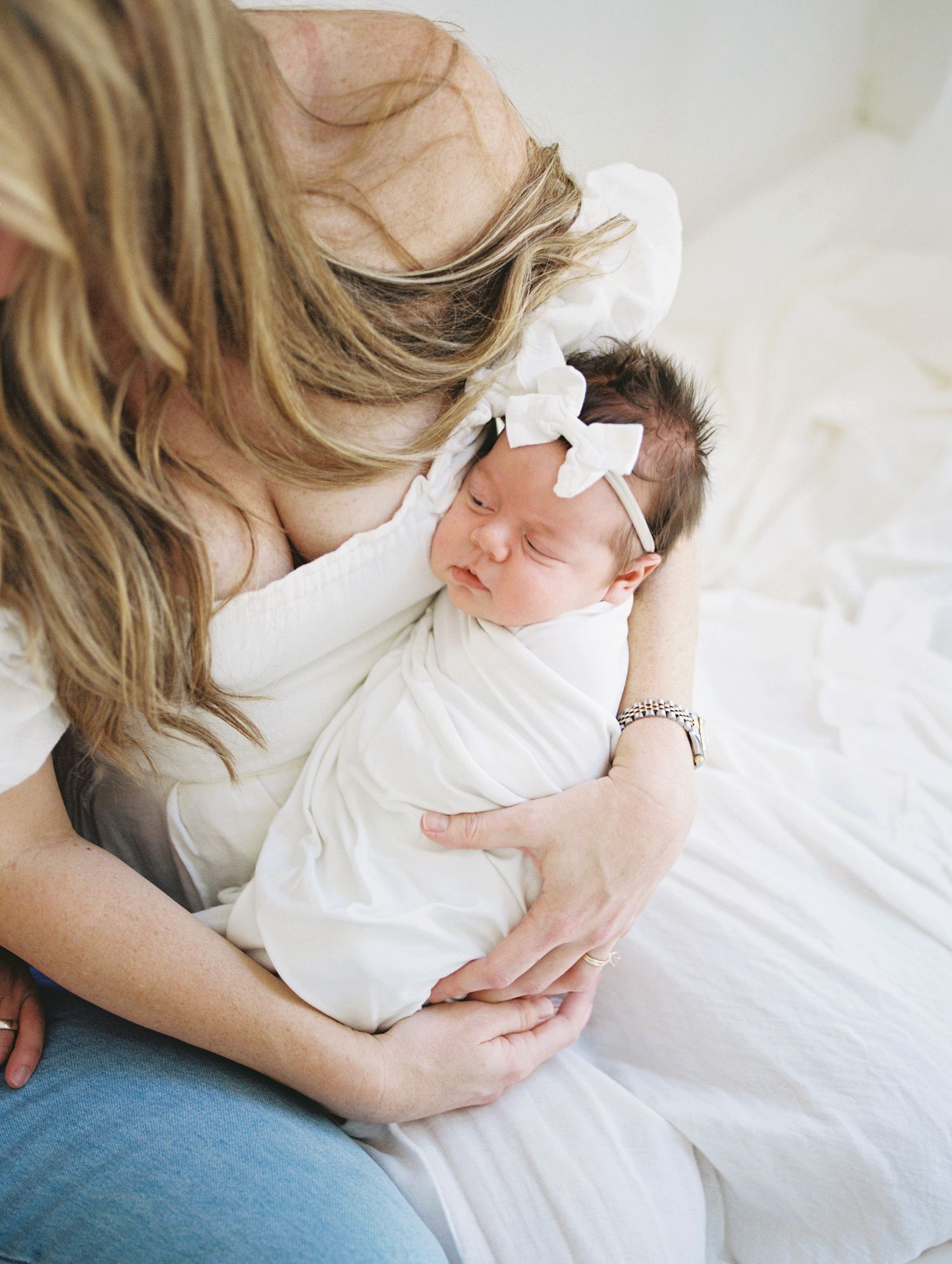 Mother with long blonde hair kissing swaddled newborn baby girl wearing white bow headband.