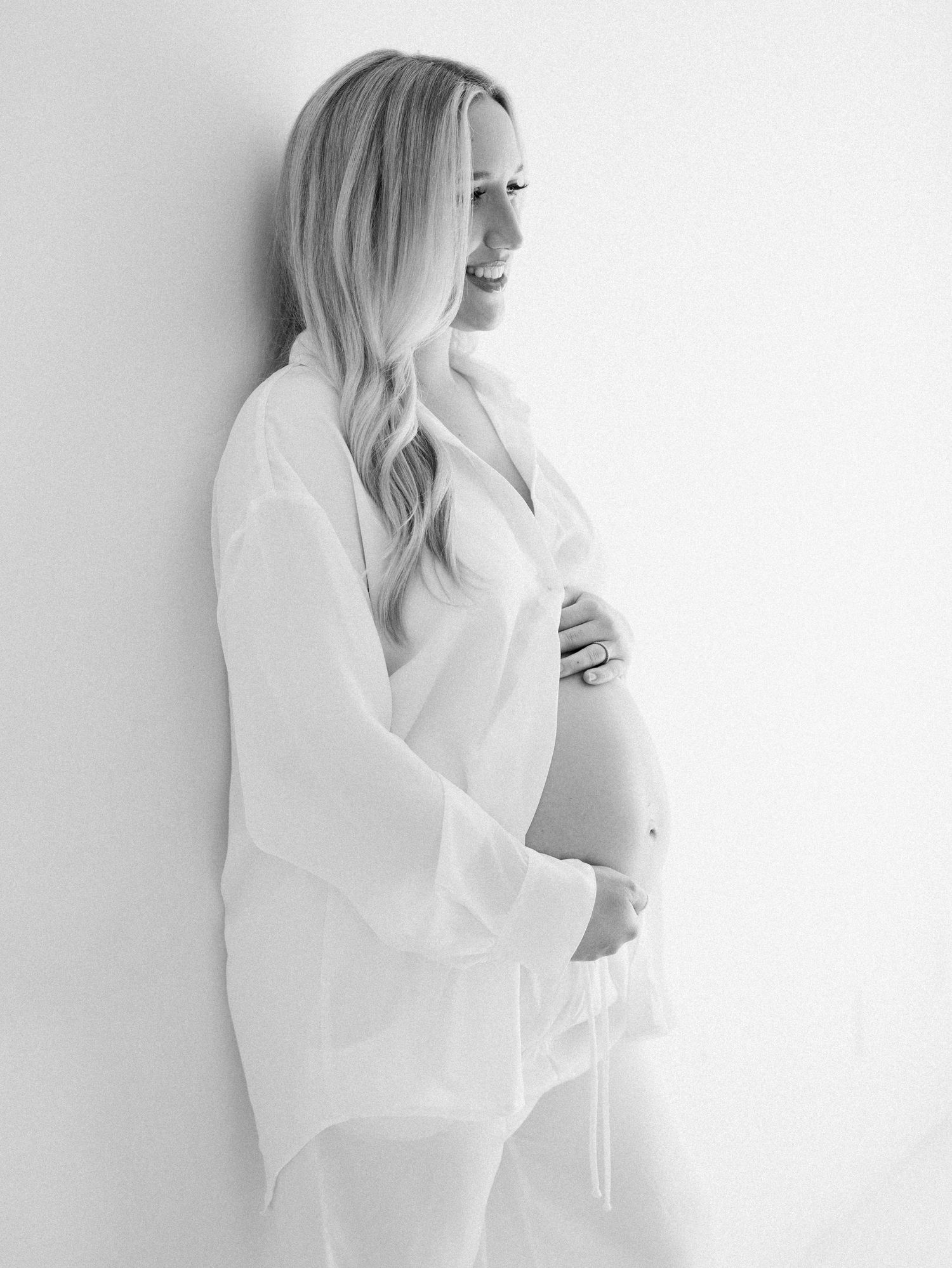 Black and white maternity photo of smiling blonde woman in white shirt cradling baby bump against white wall.