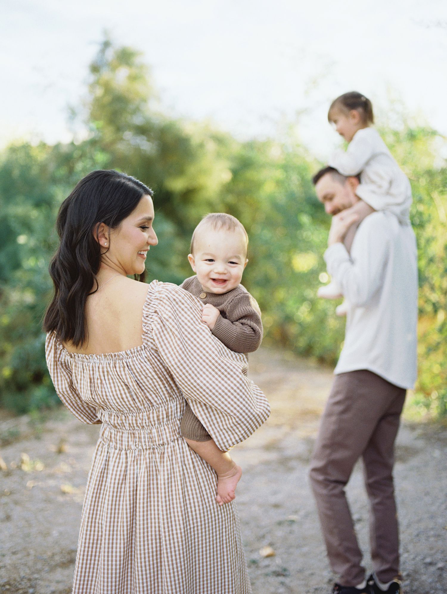 Mother holding smiling baby while father carries toddler on shoulders outdoors.