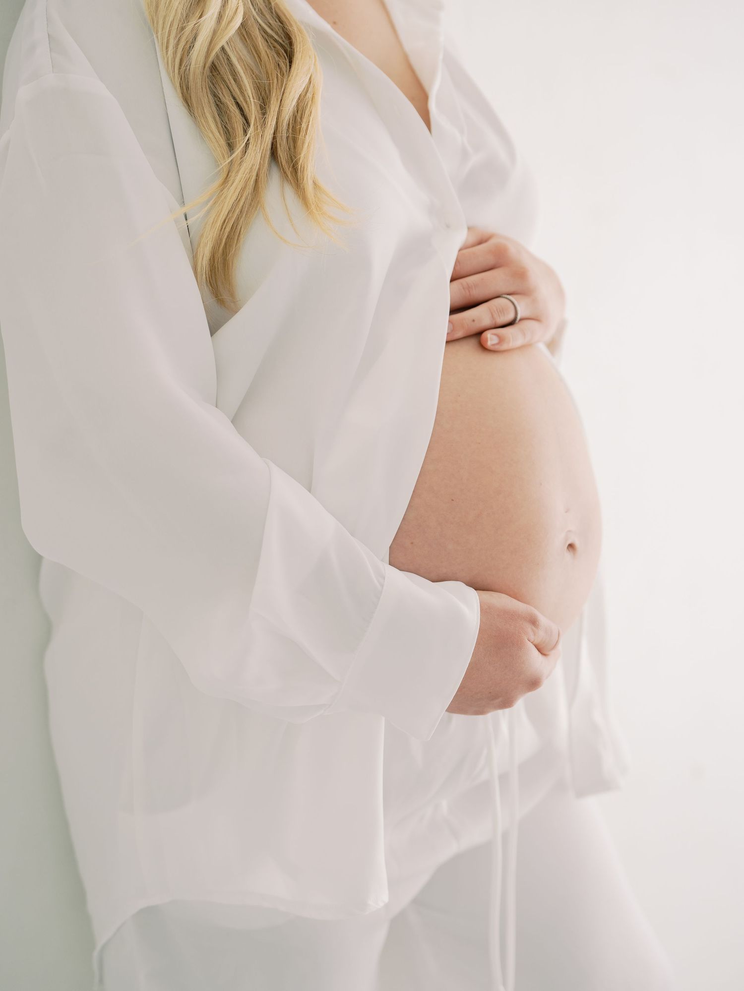 Close-up of pregnant woman in white sheer shirt gently holding bare baby bump with ringed hand.