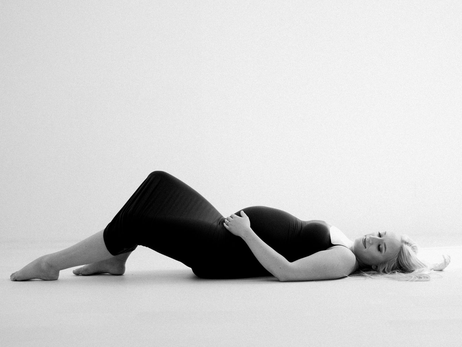 Black and white maternity photo of pregnant woman lying on floor in relaxed pose wearing dark outfit.