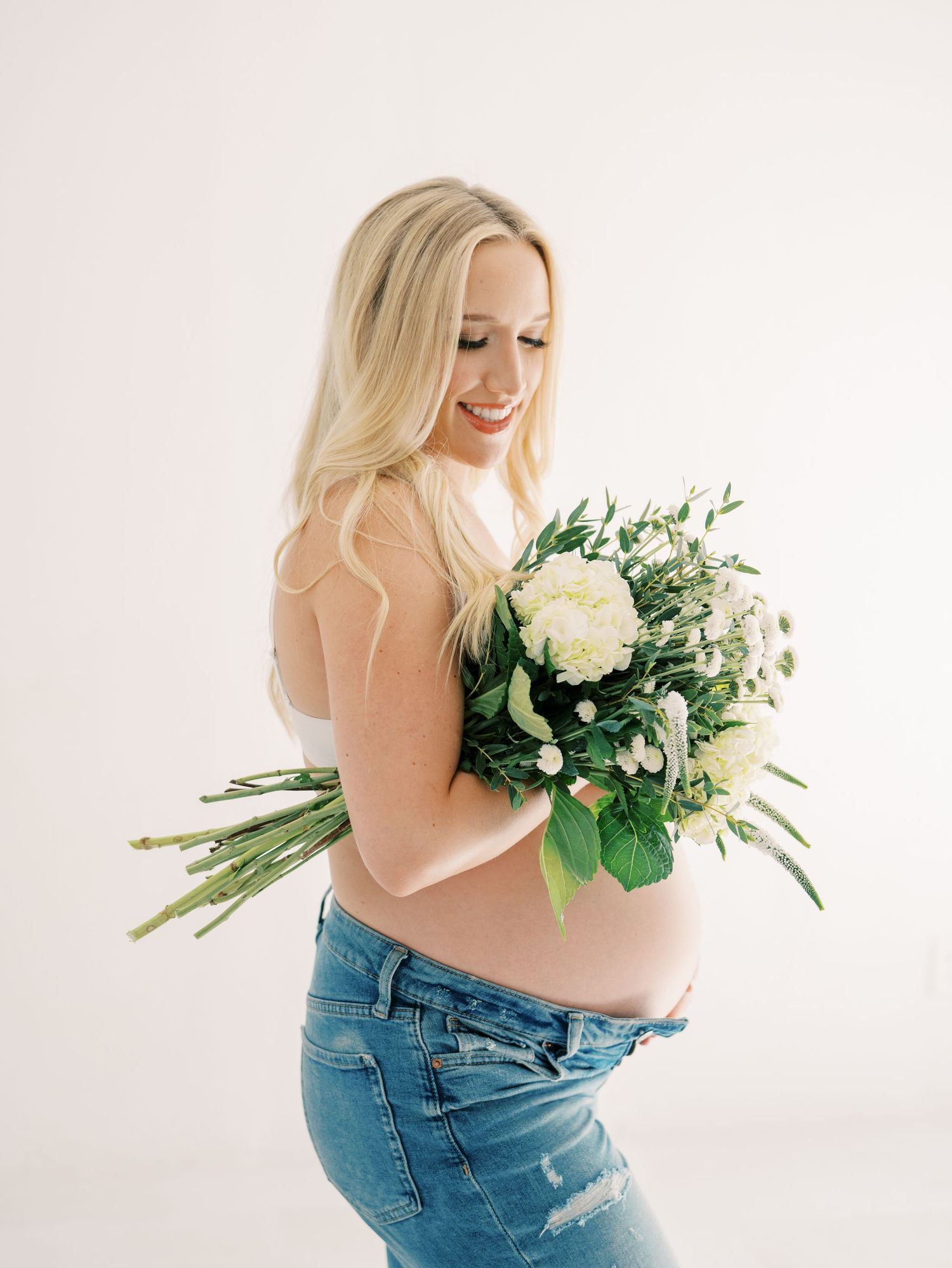 Smiling pregnant woman holding white floral bouquet in studio, wearing jeans with long blonde hair.