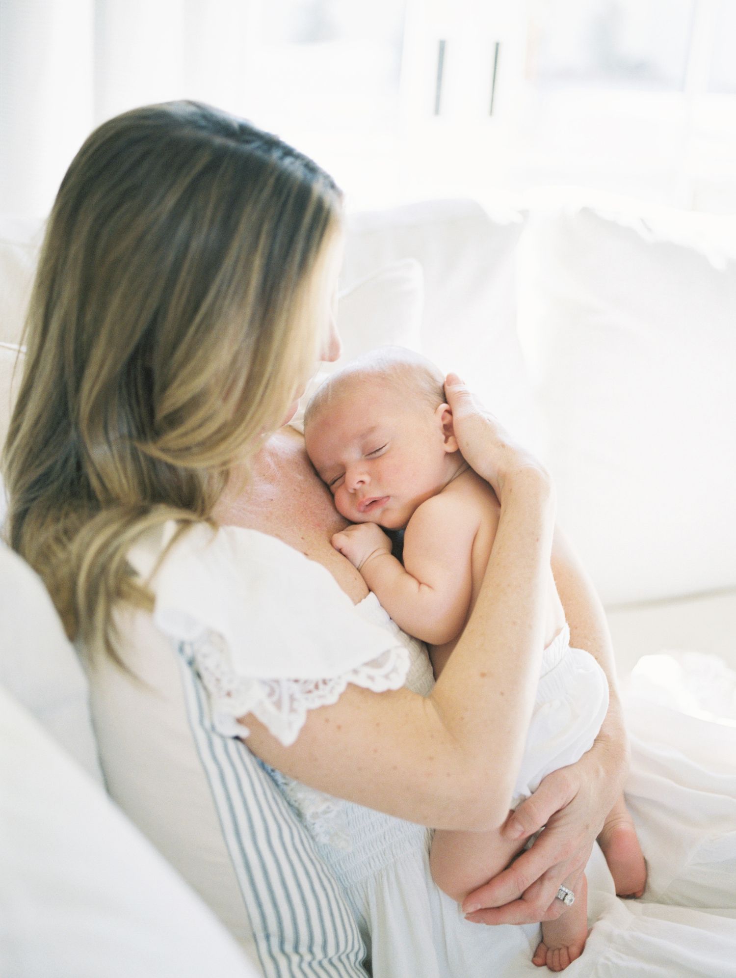 Mother in white lace dress holding sleeping newborn baby against her chest in bright white room.