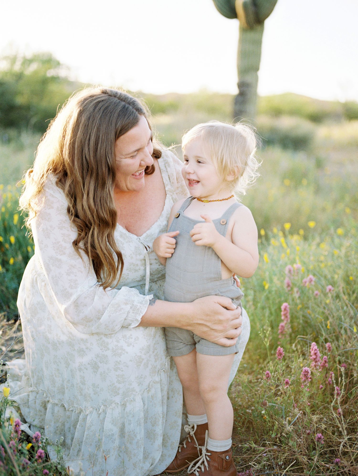 Smiling mother kneeling with happy toddler in desert wildflower field near saguaro.