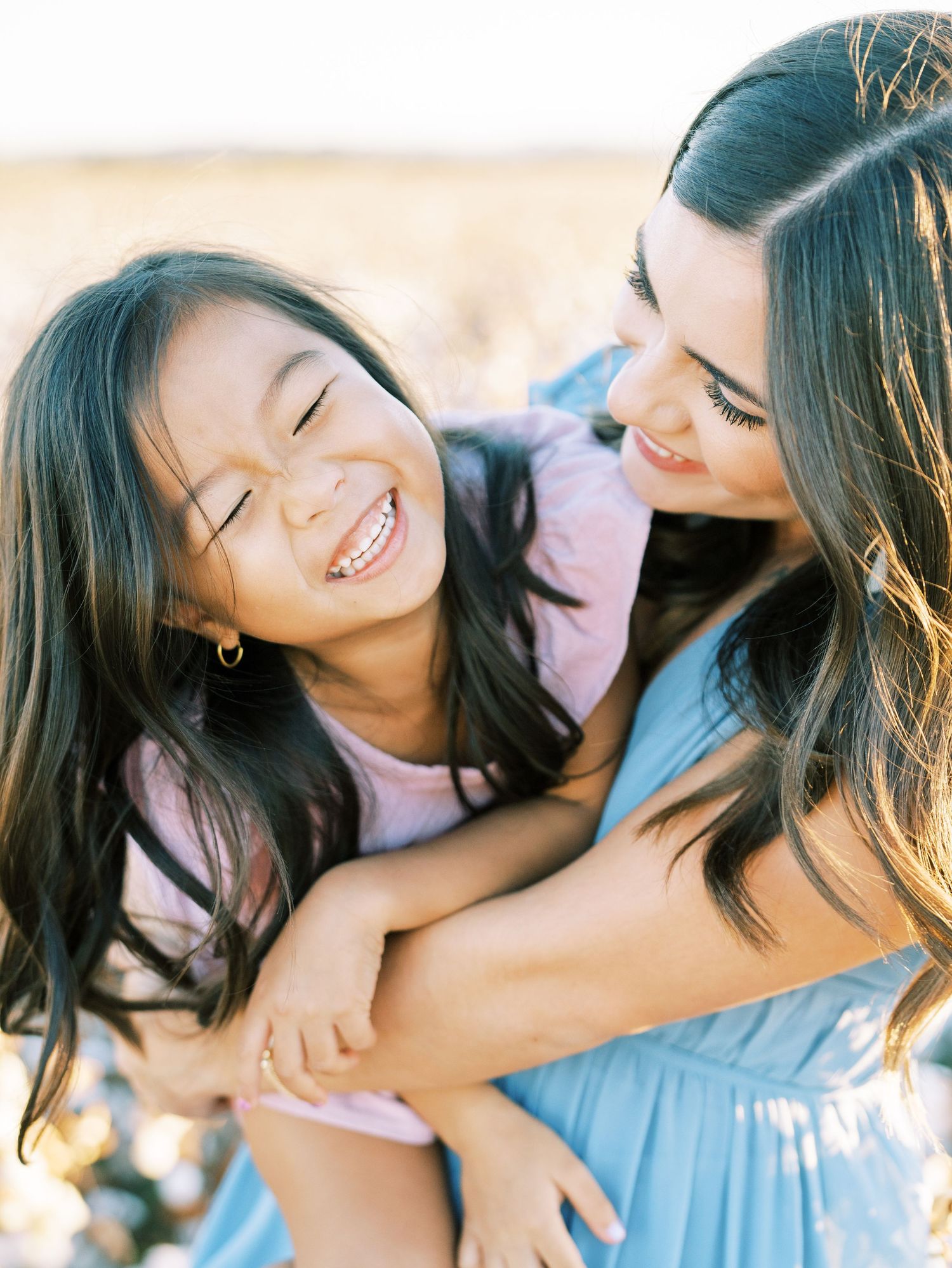 Joyful mother hugging laughing daughter in sunlit field during outdoor family portrait session.