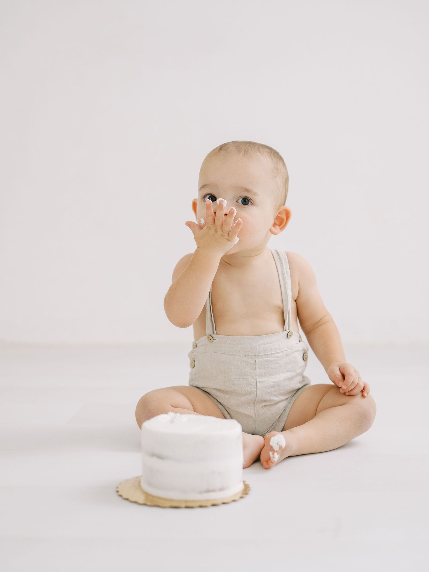 Baby boy in linen suspender shorts covering mouth with hand beside first birthday smash cake.