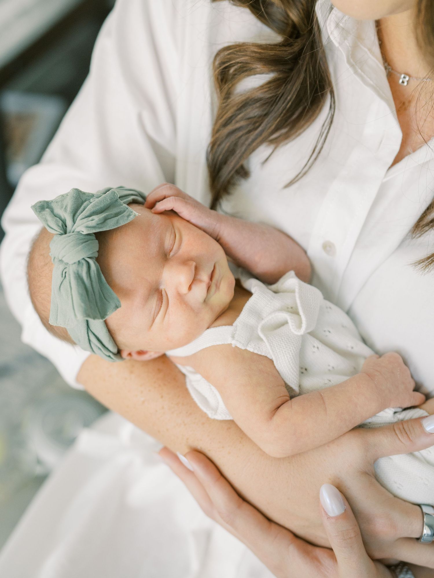 Mother in white shirt tenderly holding sleeping newborn baby girl with sage green bow headband.