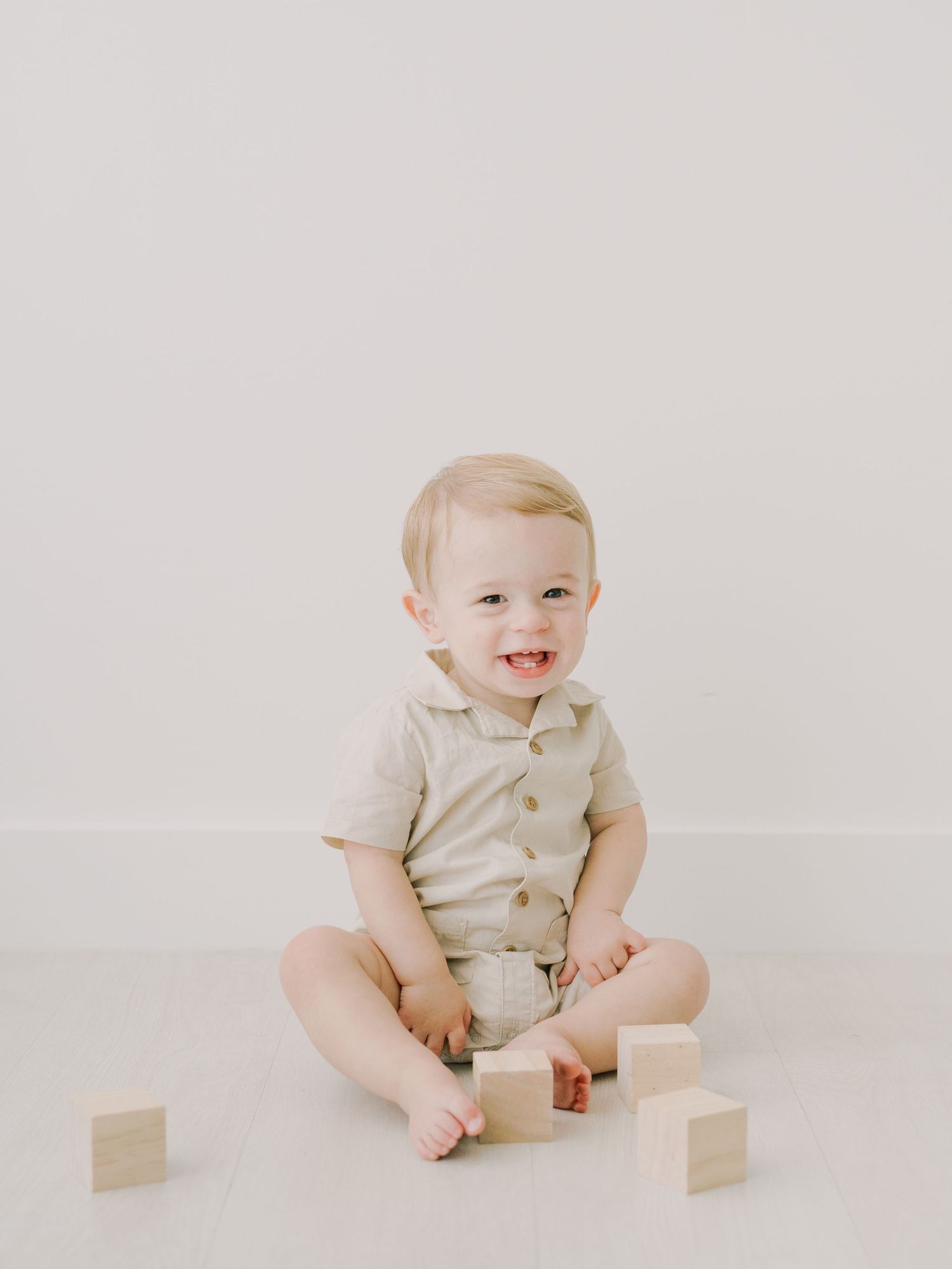 Adorable smiling baby boy sitting on white floor in beige outfit with wooden blocks nearby.