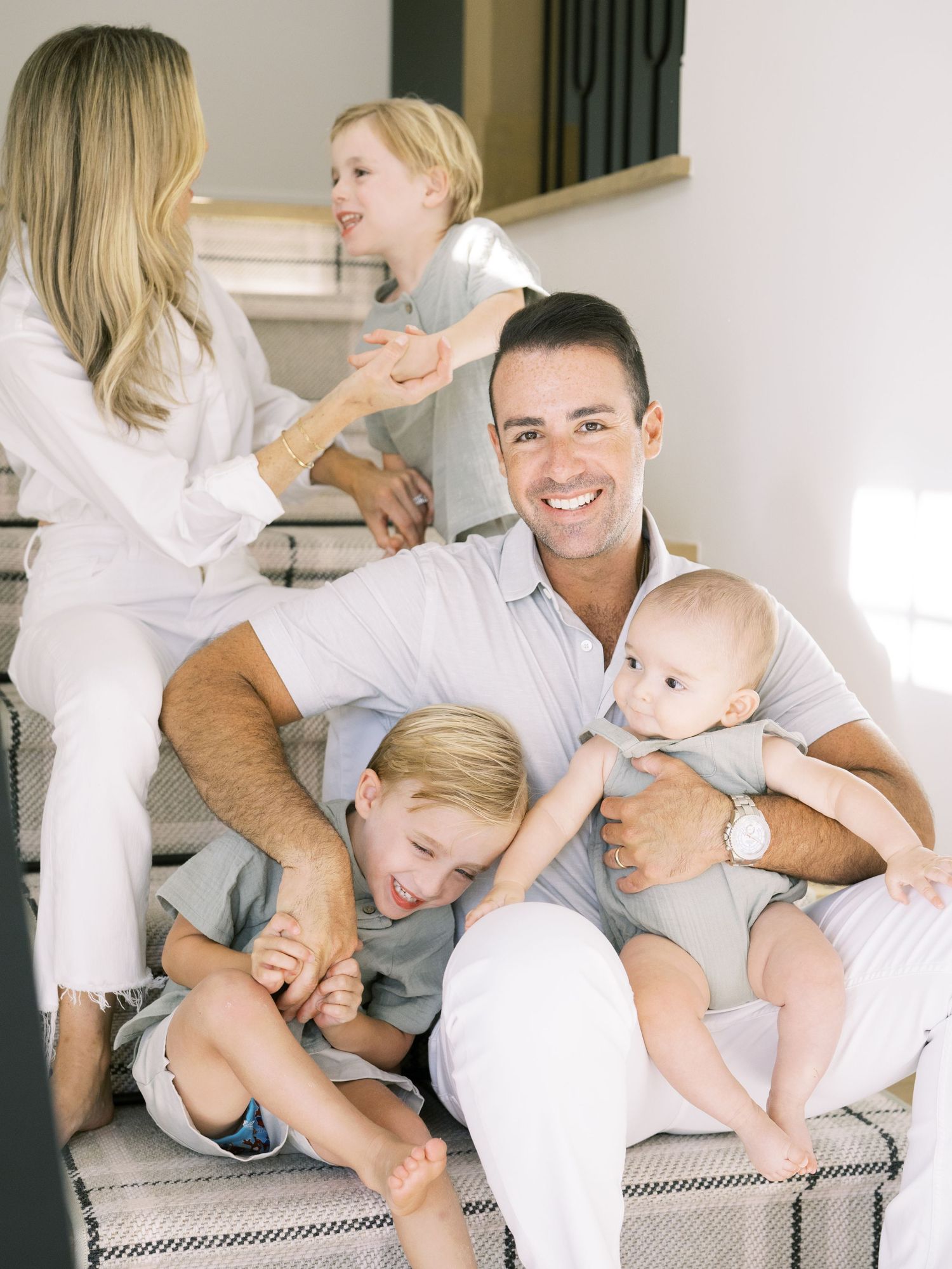 A dad smiles at the camera while holding a baby as two boys playfully climb on him on stairs.
