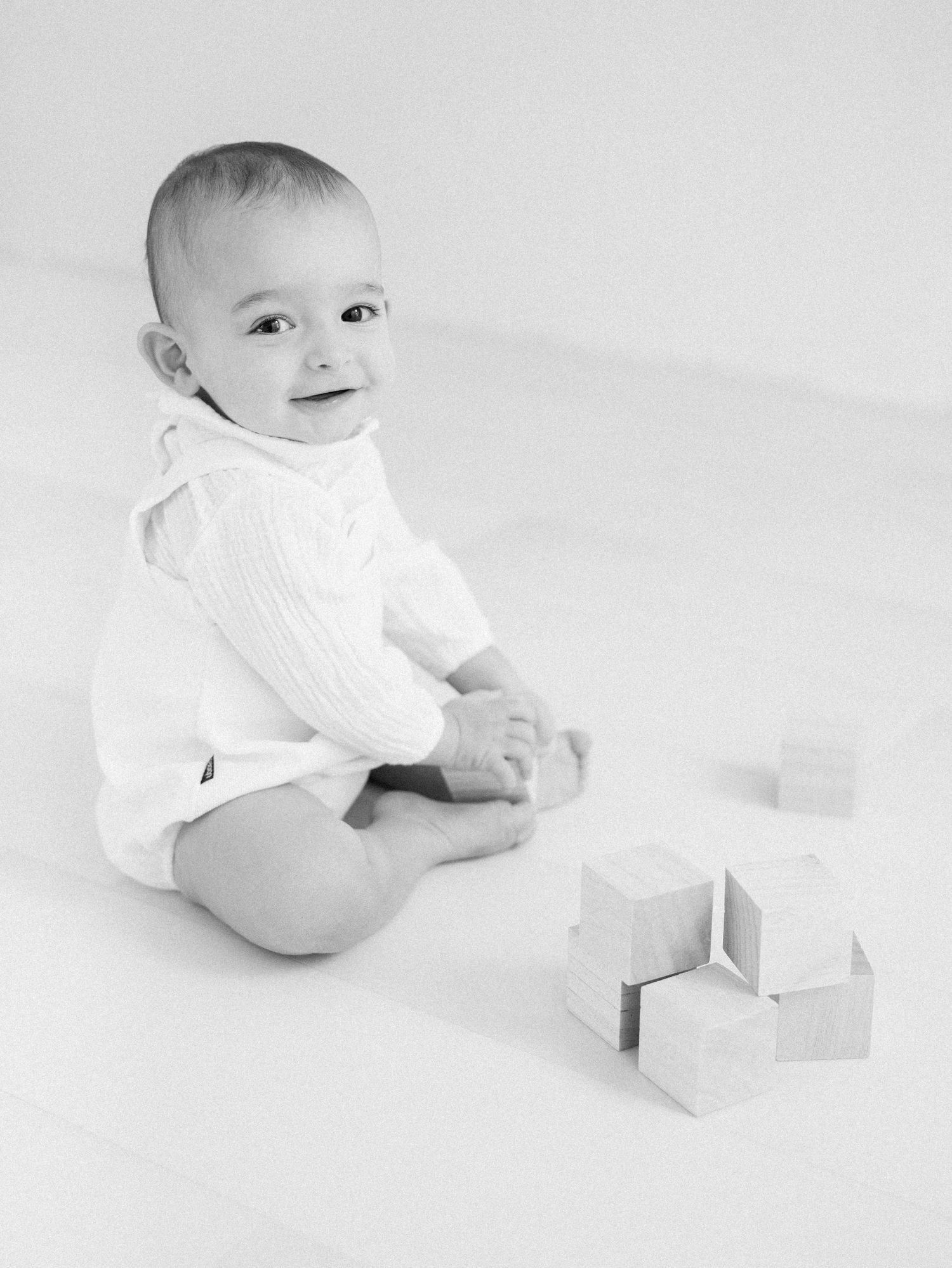 Black and white portrait of smiling baby in white knit outfit sitting with wooden blocks.