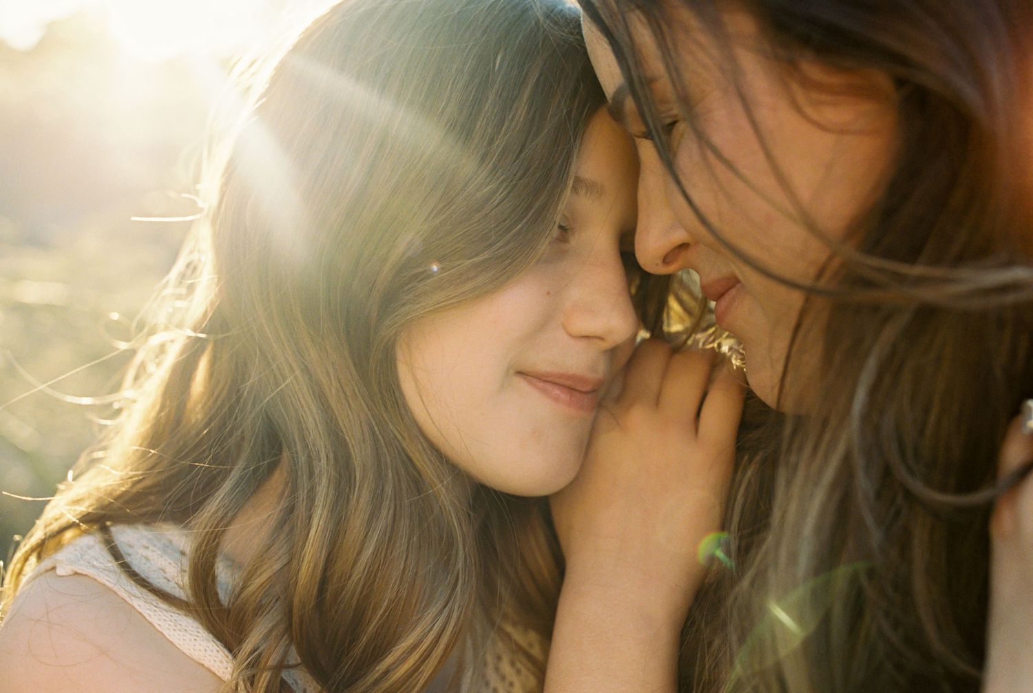 Mother and daughter sharing a tender forehead-to-forehead moment in golden sunlight outdoors.