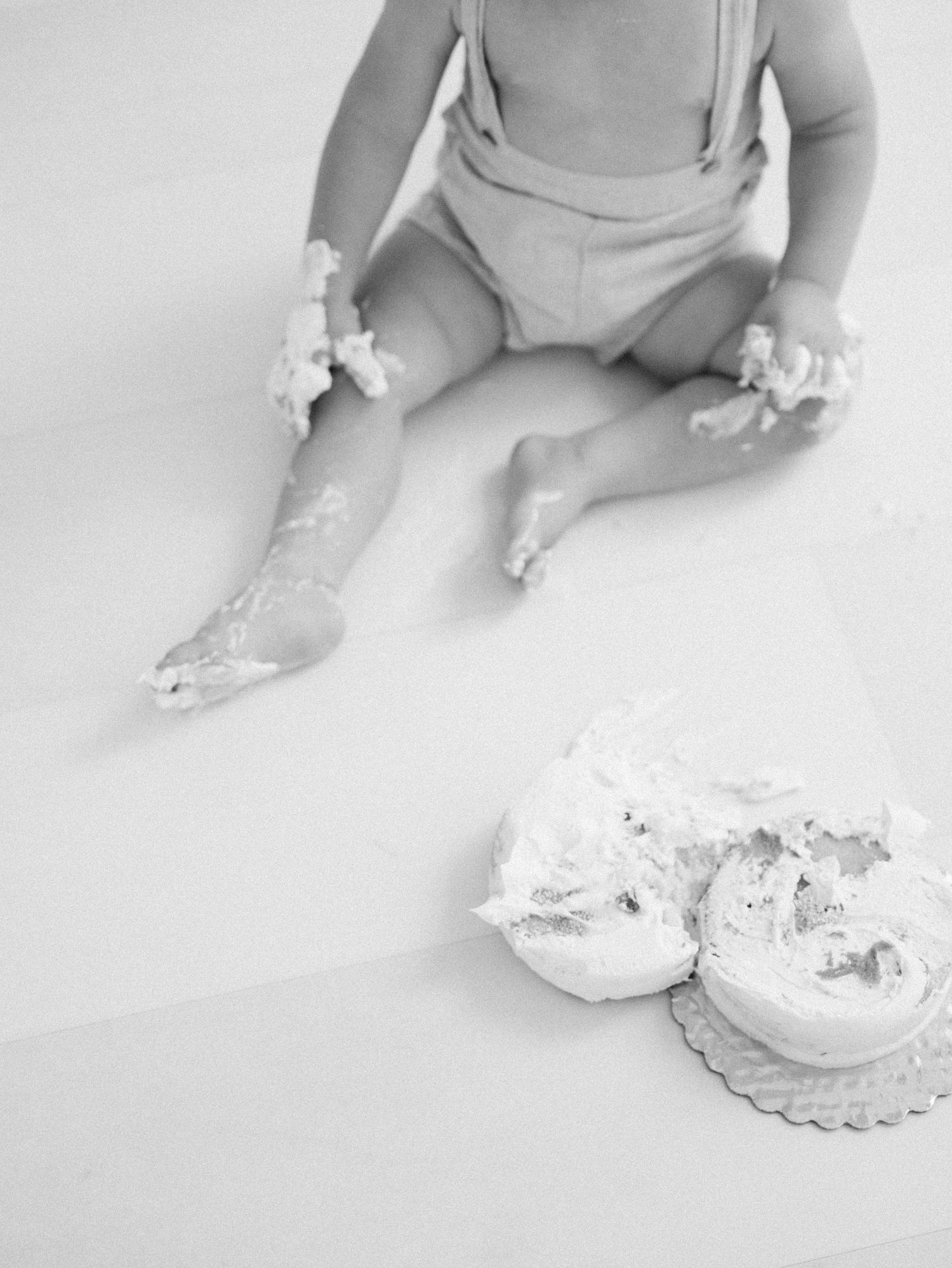 Black and white photo of baby sitting with smashed birthday cake on legs.