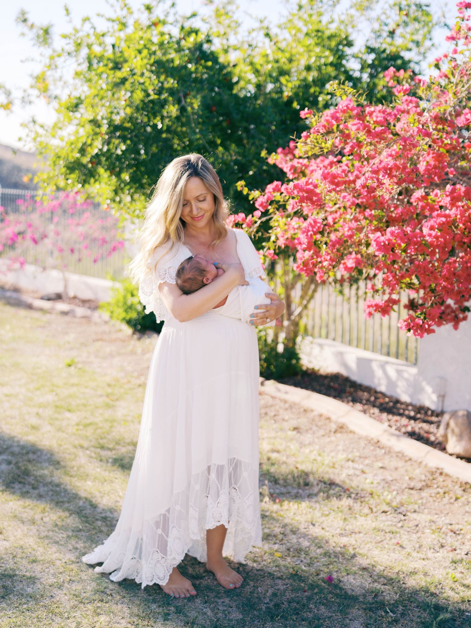 New mother in white lace maxi dress cradling newborn baby outdoors near blooming pink flowering tree.