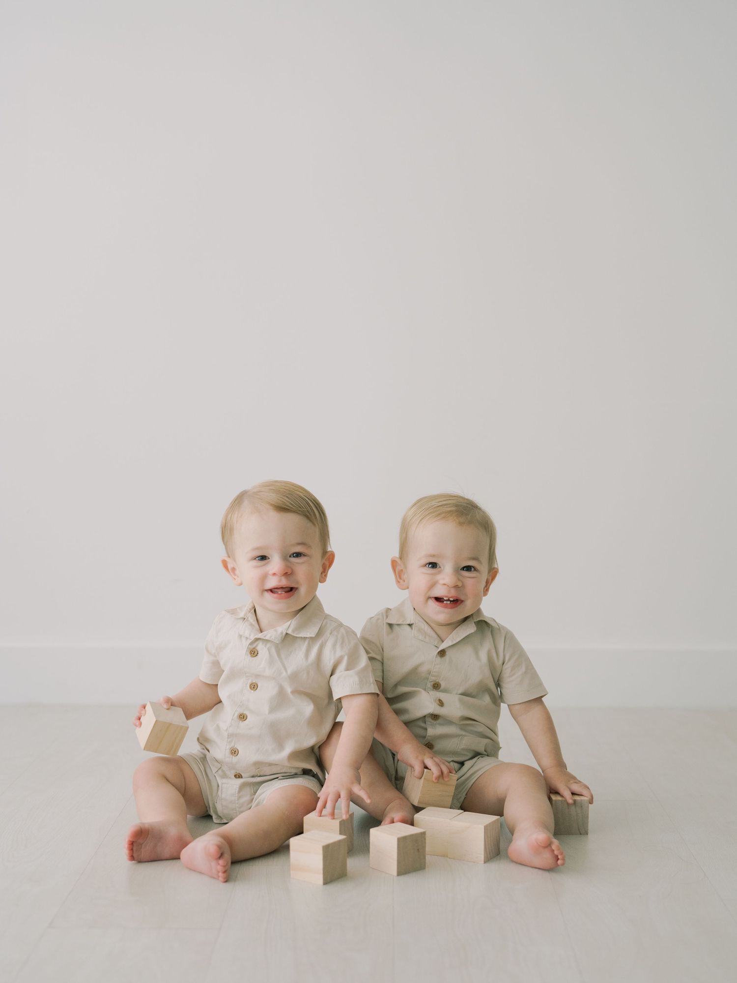 Two smiling baby twins sitting on white floor in matching beige outfits playing with wooden blocks.