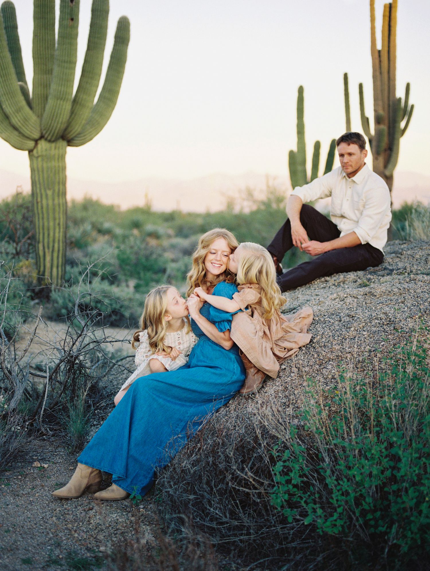 Mother hugging two daughters on boulder with saguaro cacti at desert sunset.