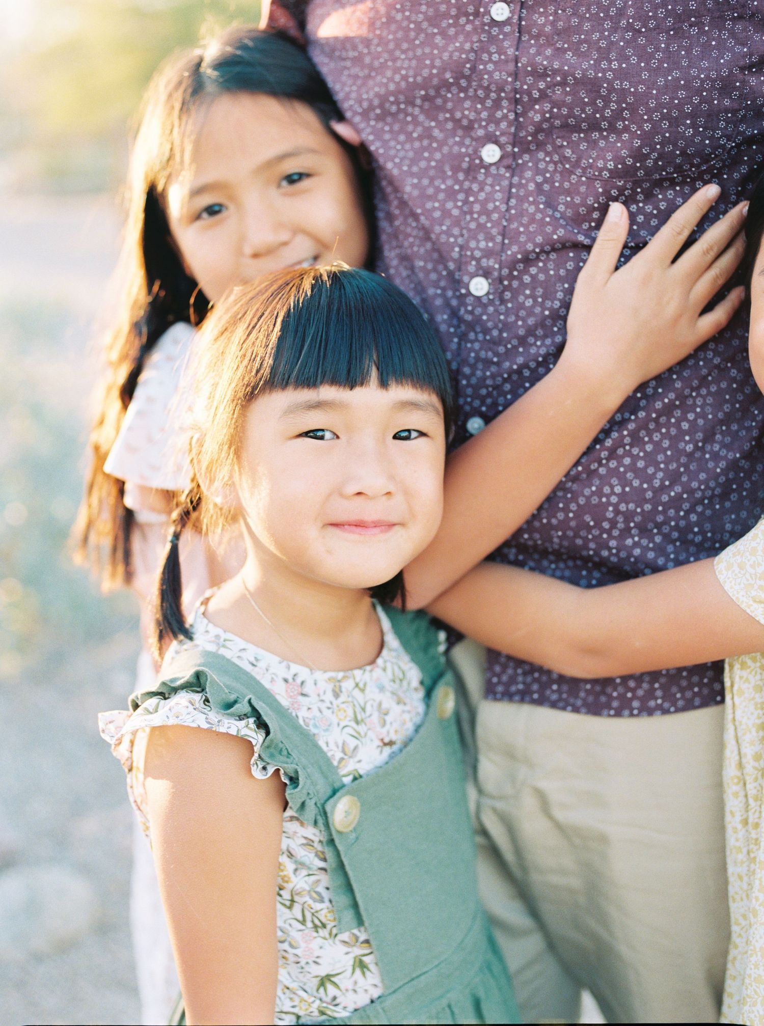 Two young Asian girls with bangs smile warmly while hugging a parent in golden afternoon light.