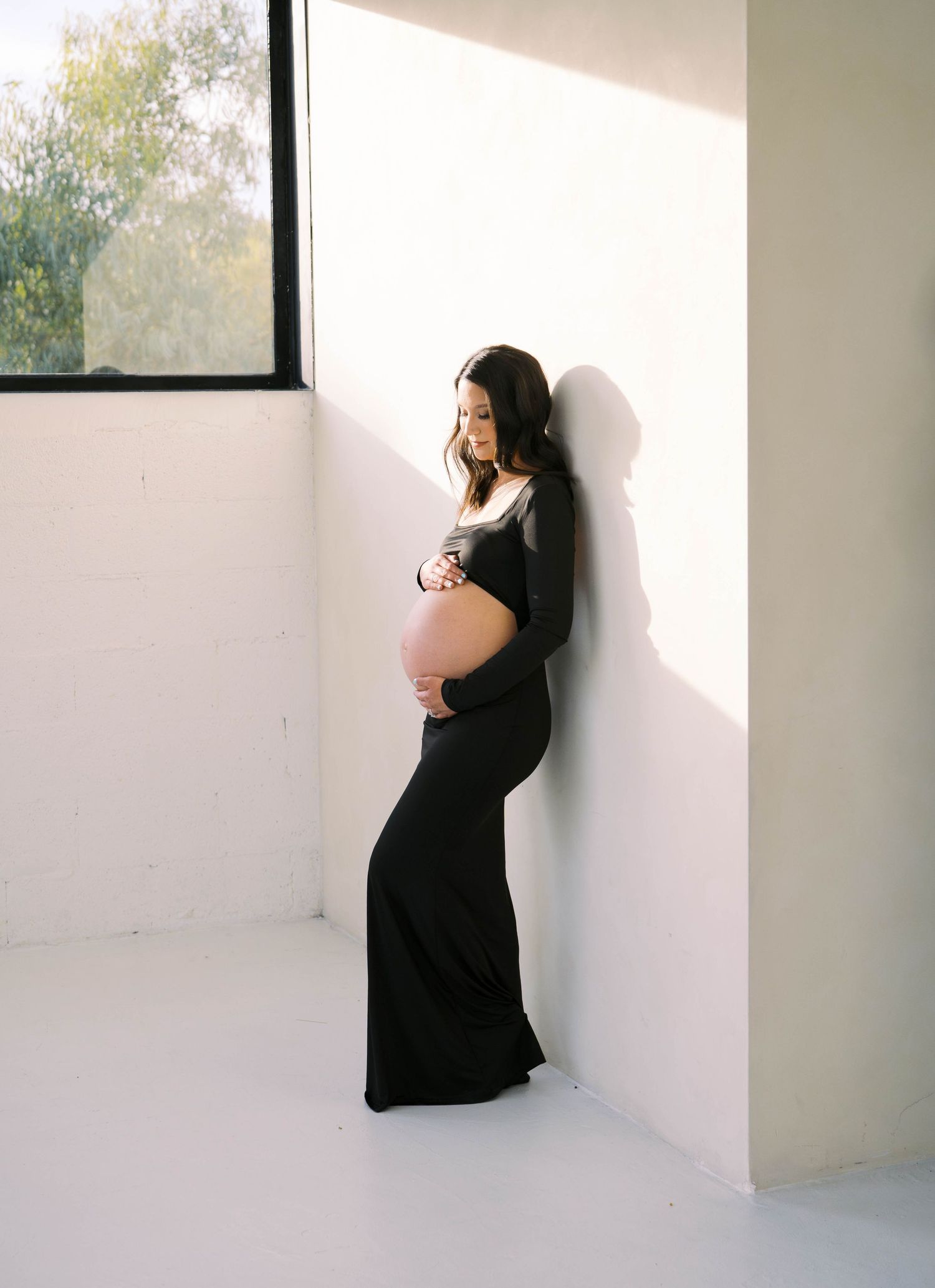 A pregnant woman in a black form-fitting gown leans against a sunlit white wall holding her baby bump.