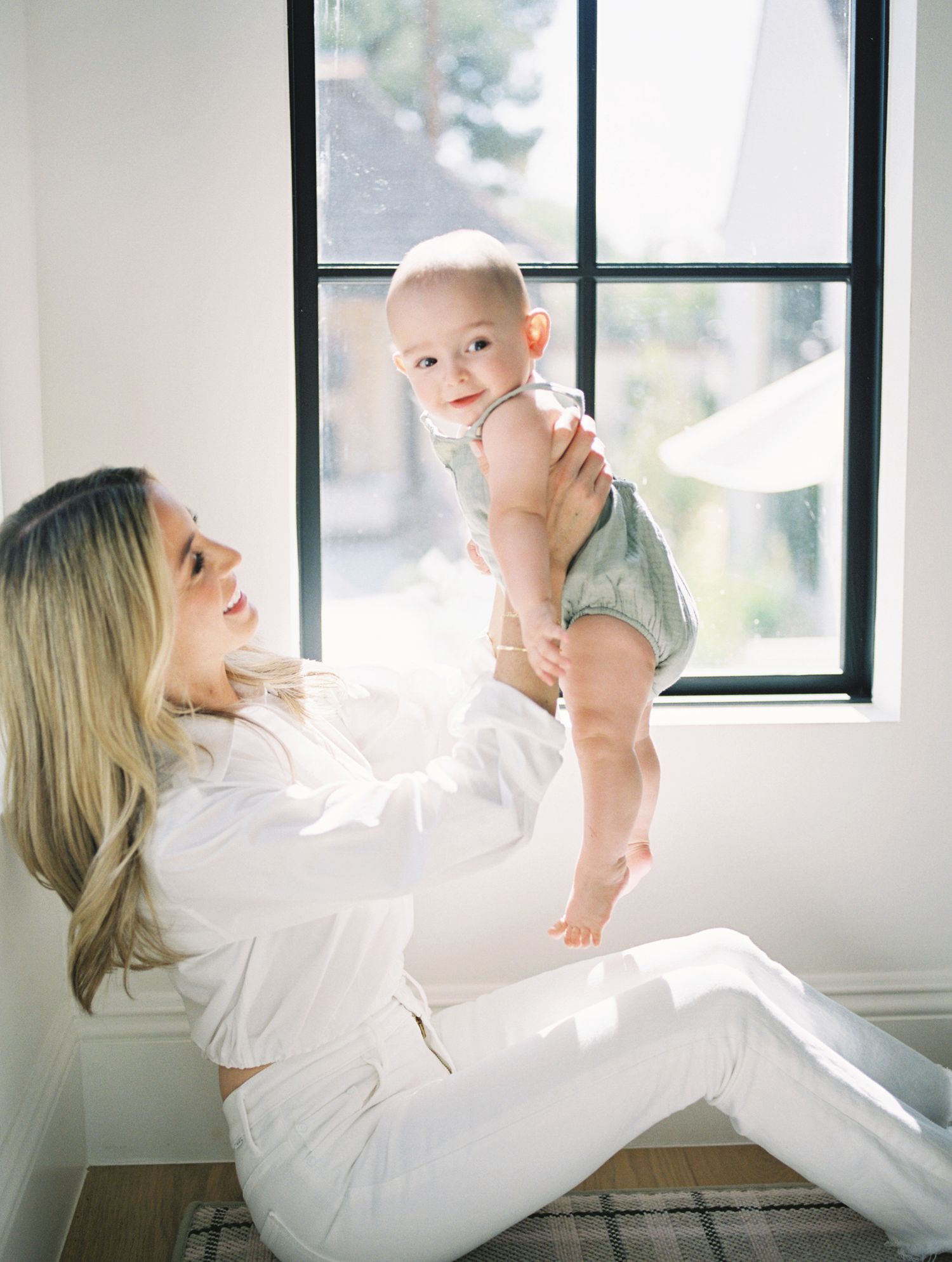 Blonde mother sitting by window lifting smiling baby wearing green romper in bright white room.
