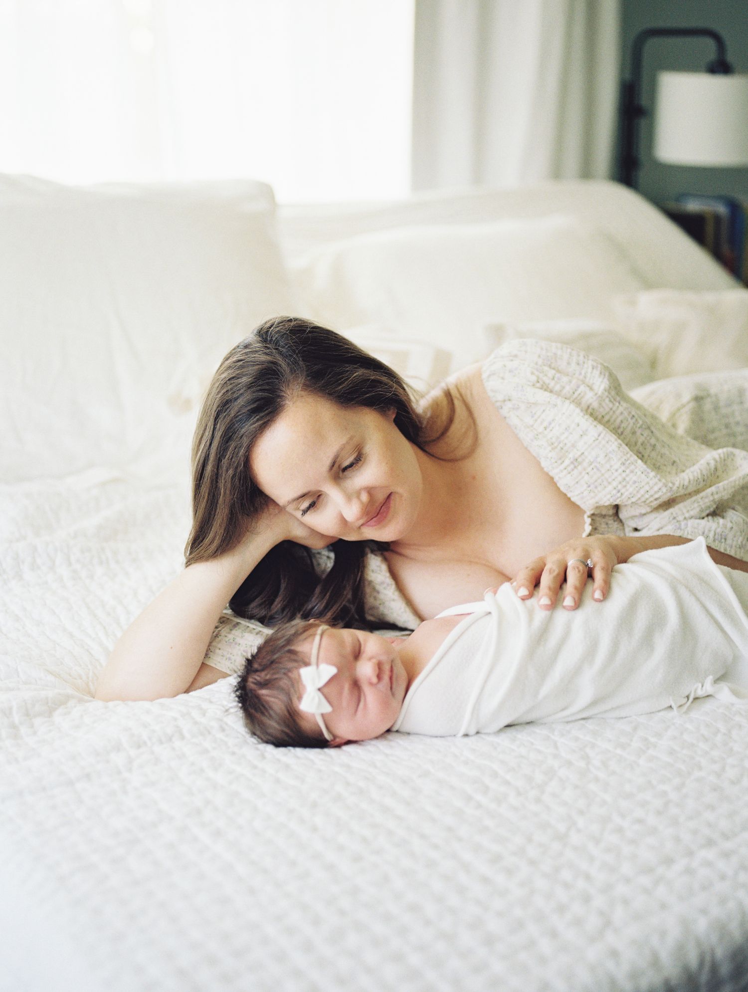 Brunette mother lying on white bed smiling tenderly at swaddled newborn baby with pink bow headband.