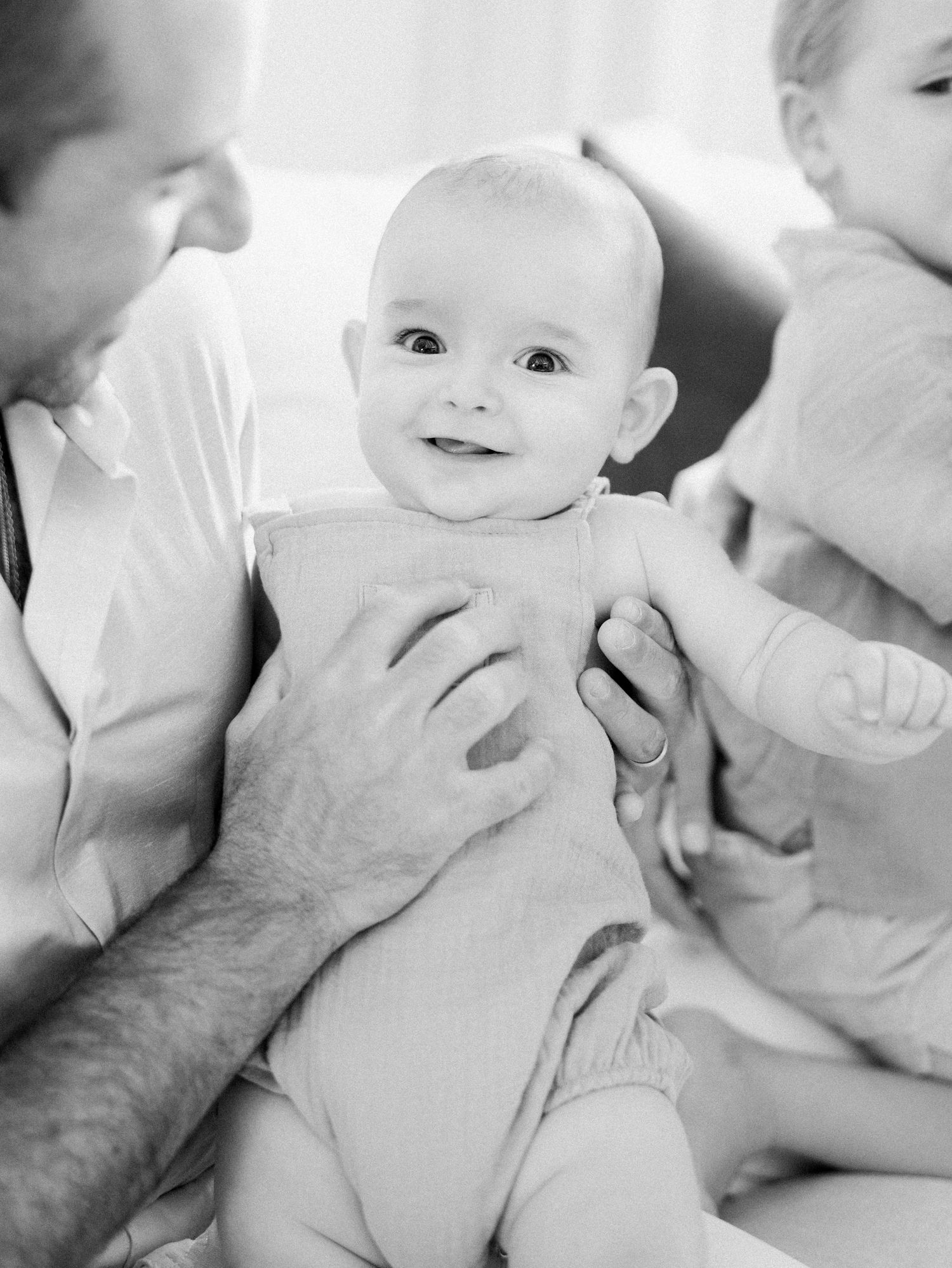A smiling baby is held lovingly by parents in a soft black and white family portrait.