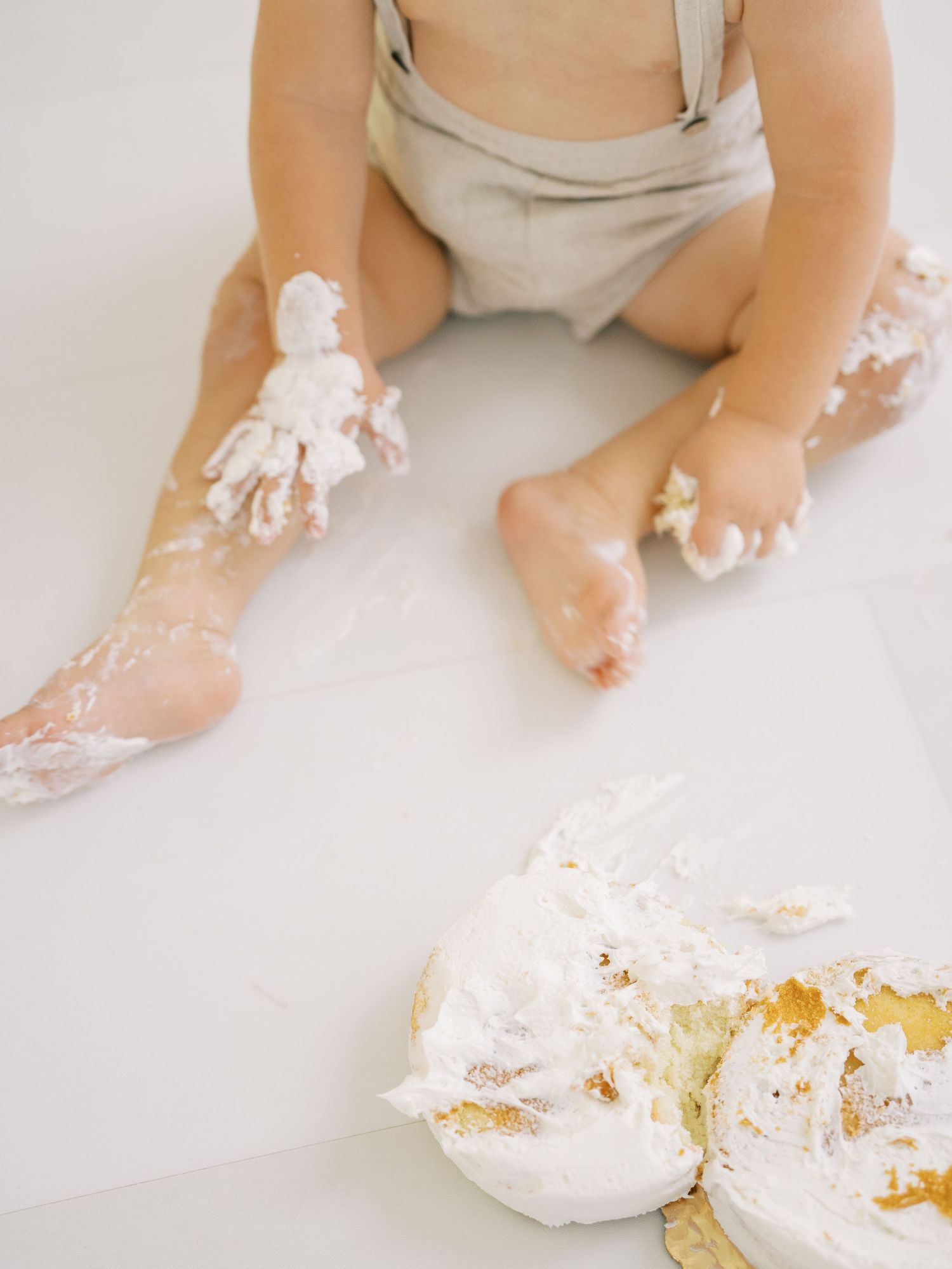 Close-up of baby's frosting-covered feet and hands beside smashed white birthday cake on floor.
