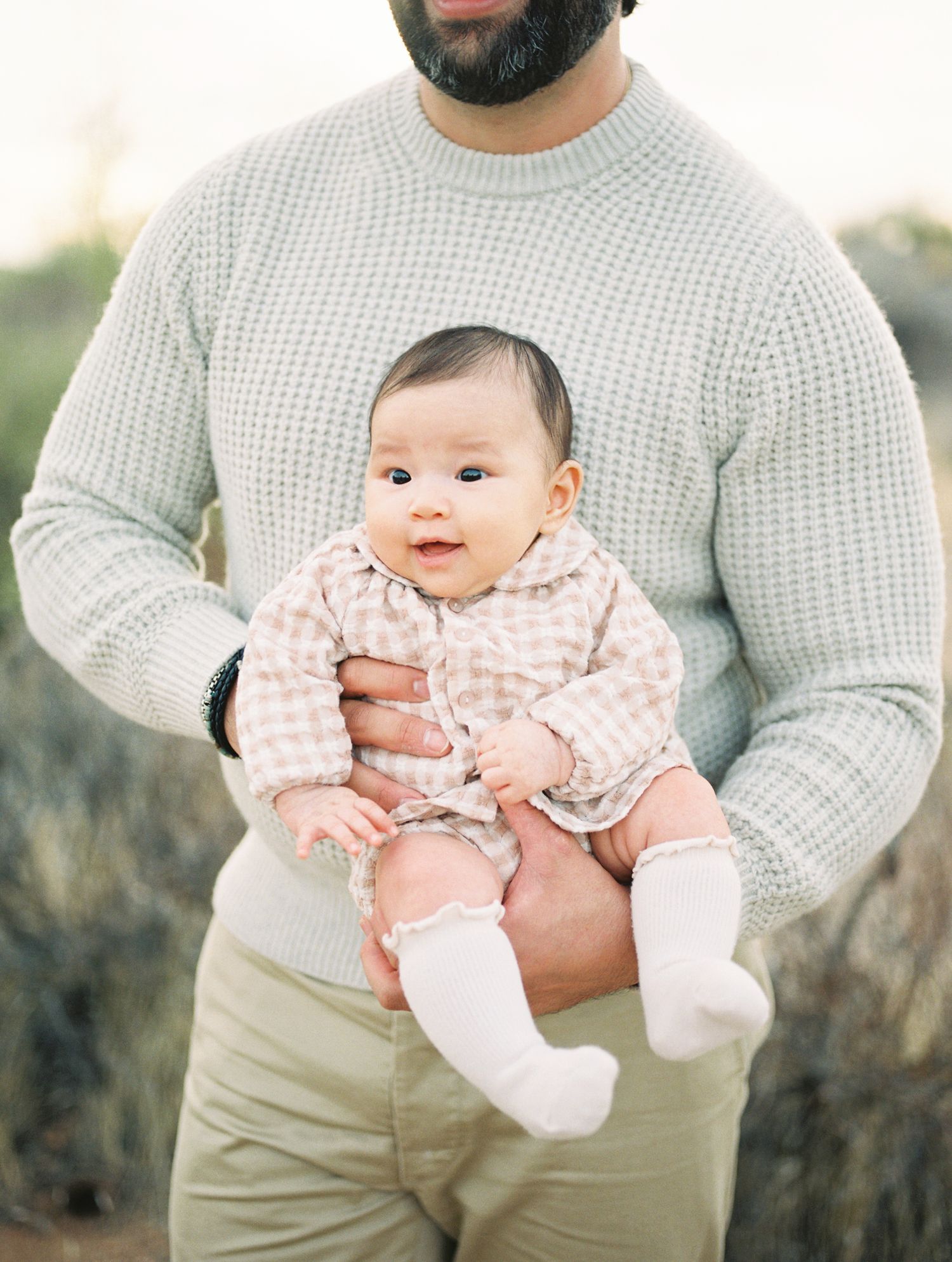 Bearded father in mint sweater holding smiling baby girl in pink gingham outfit outdoors.