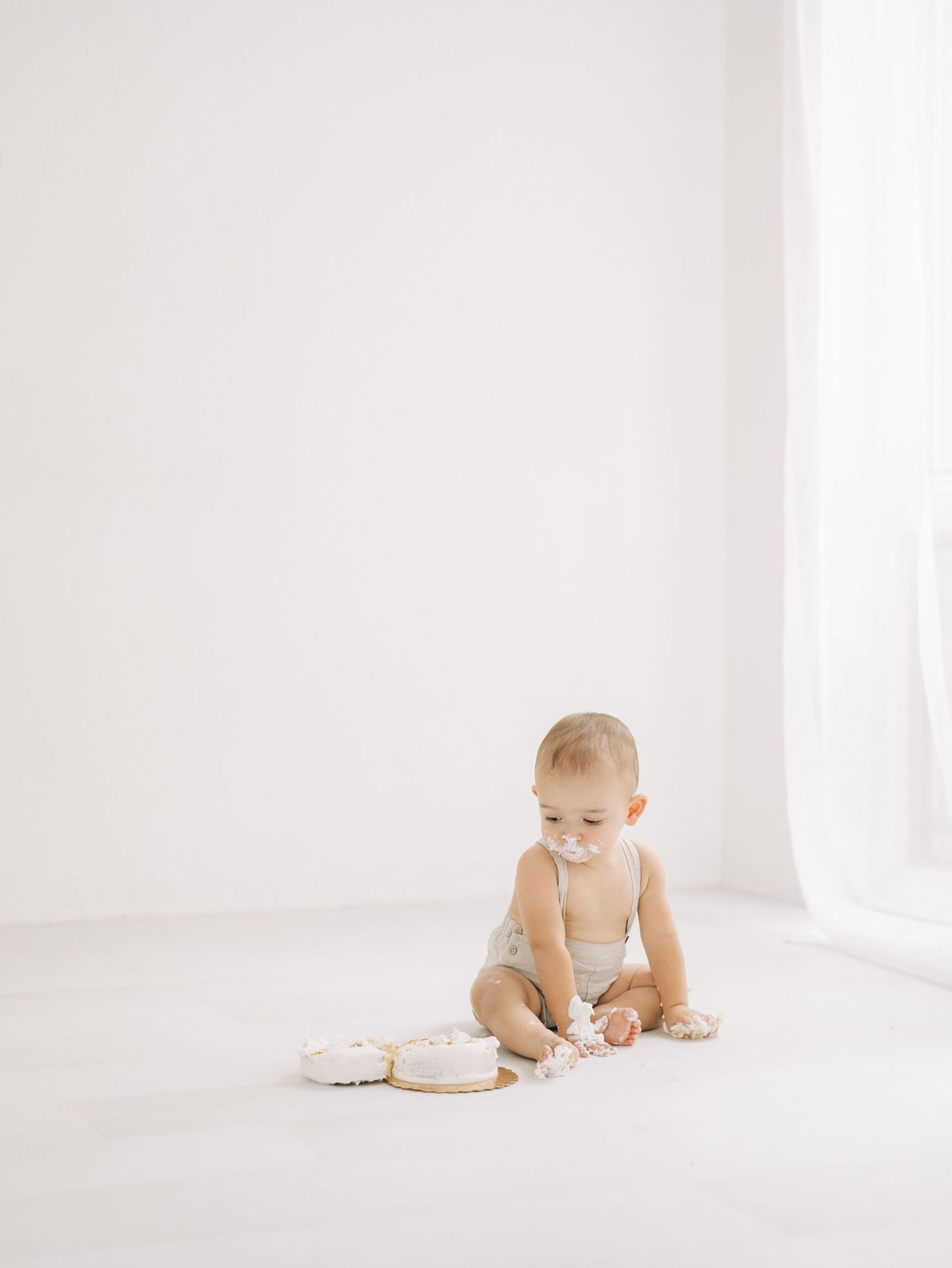 Baby in neutral romper exploring smash cake in bright white studio during first birthday session.