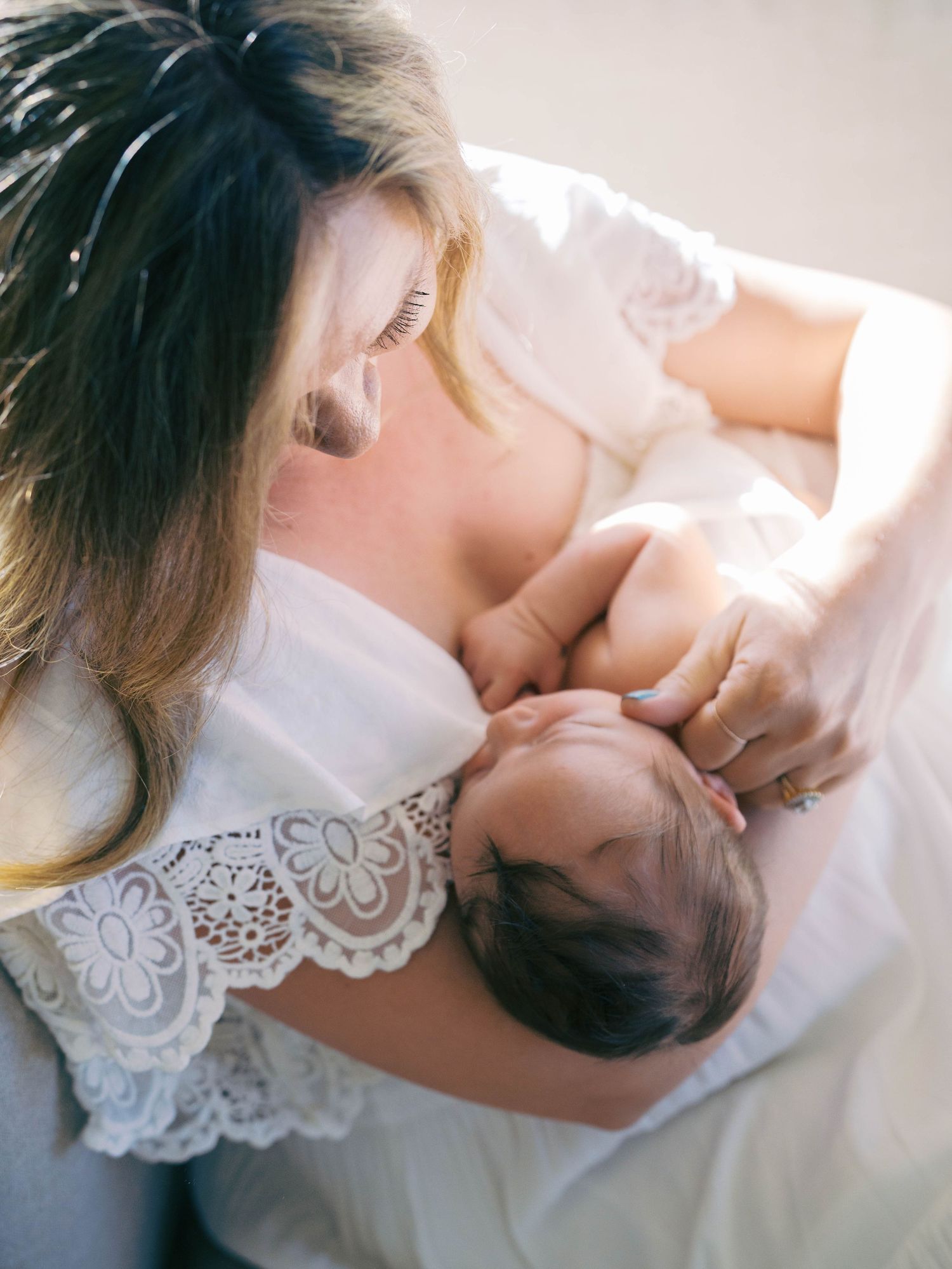 Mother in white lace dress tenderly kissing her sleeping newborn baby from above.
