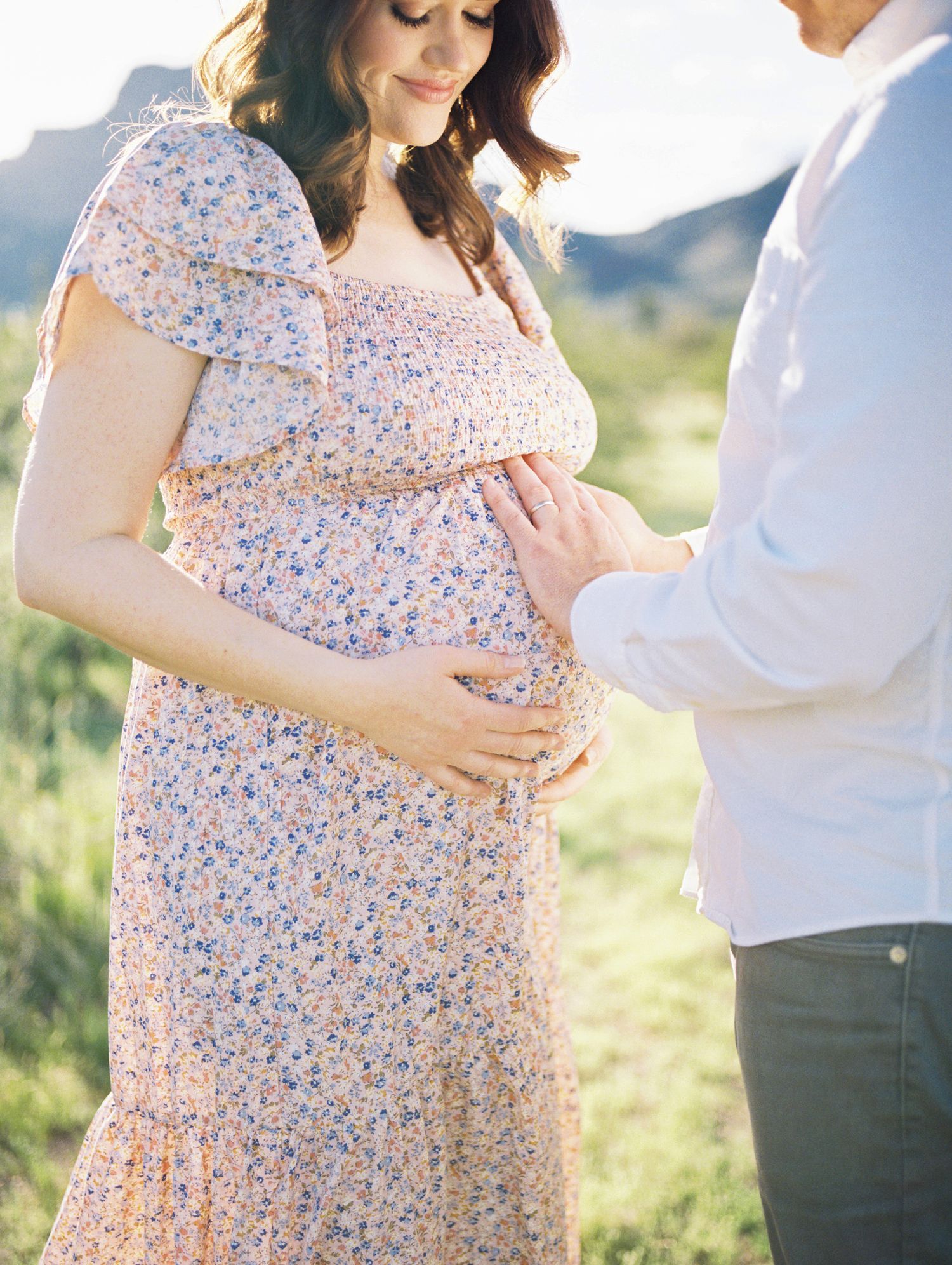A pregnant woman in a floral dress and her partner tenderly hold her baby bump in a scenic outdoor setting.