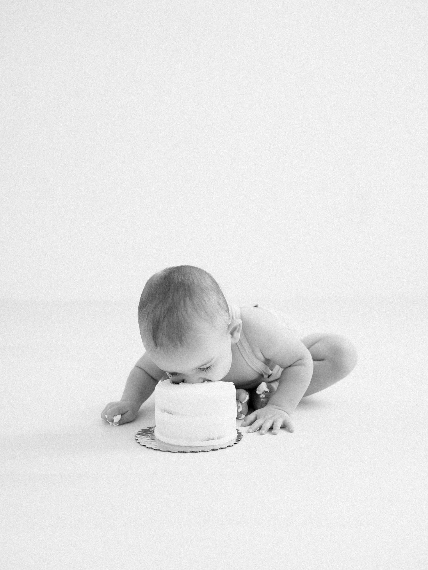 Black and white photo of baby eating first birthday cake on white studio floor.