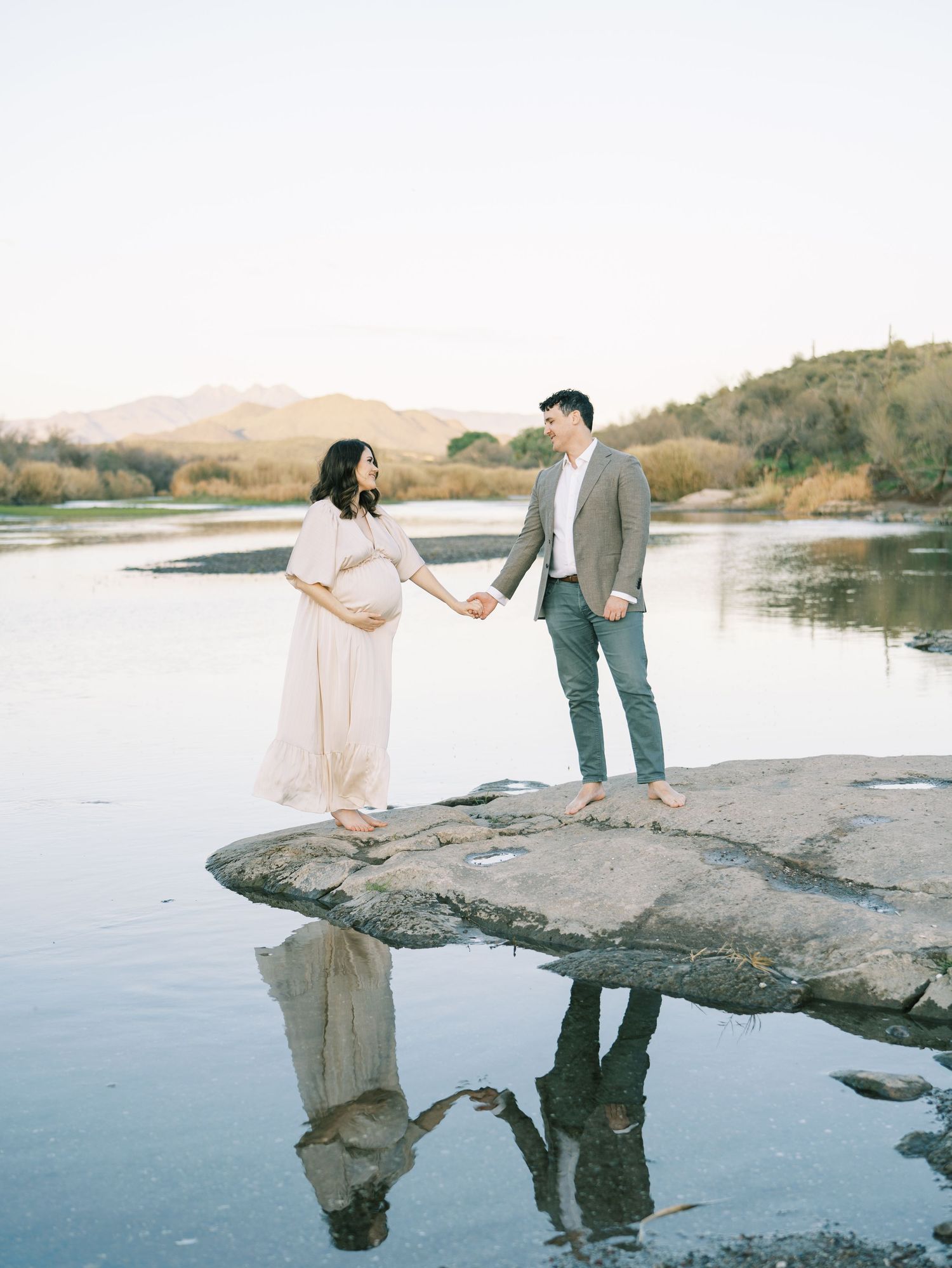 A pregnant couple holds hands on a reflective rock surface with stunning mountain and lake scenery behind them.