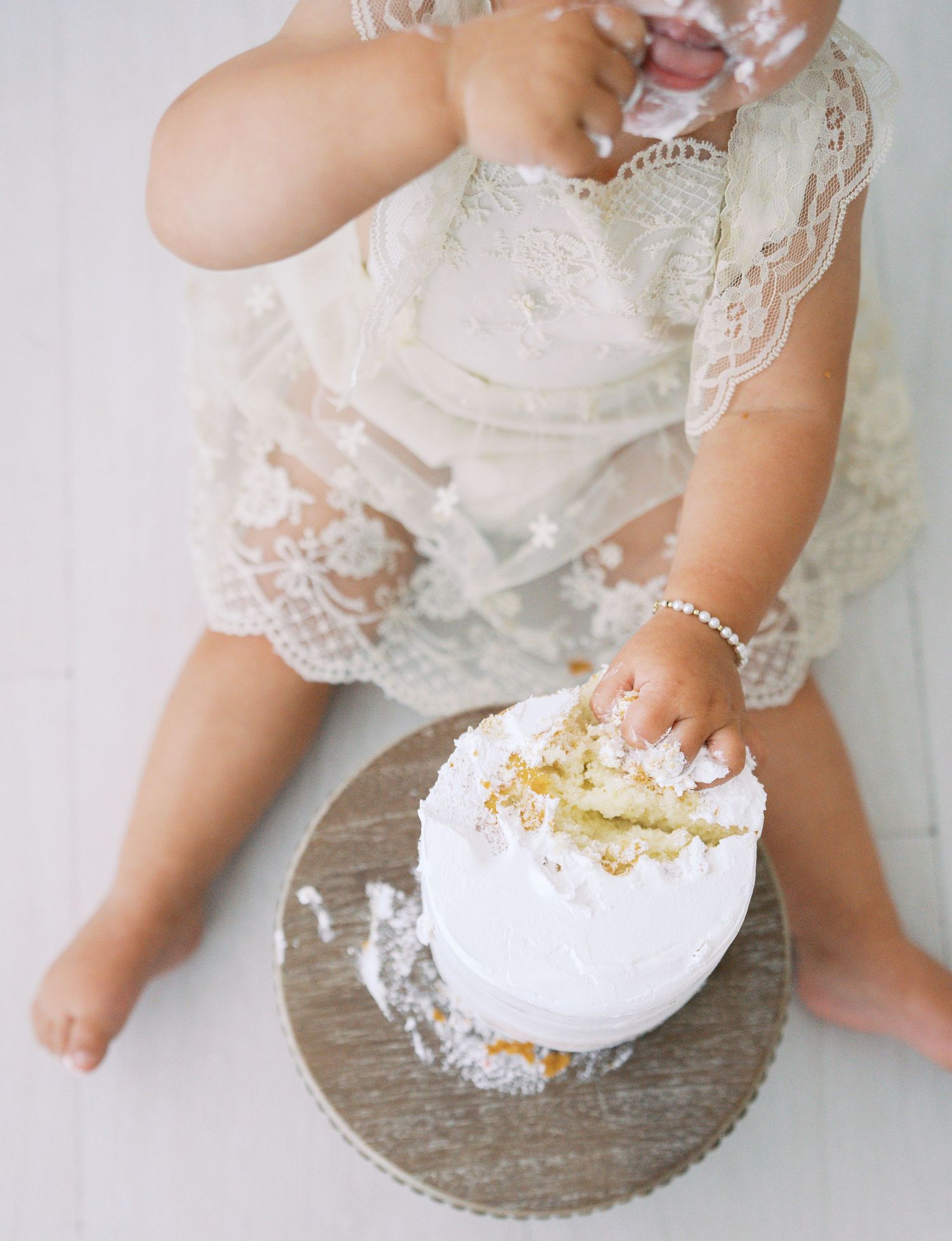 Baby girl in lace romper smashing white first birthday cake on wooden stand in bright studio.
