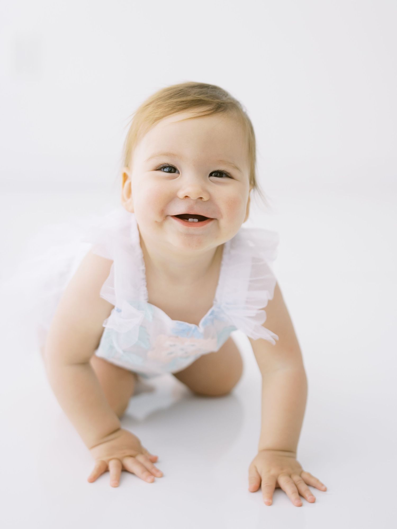 Happy baby girl in white ruffled romper crawling and smiling on bright white background.