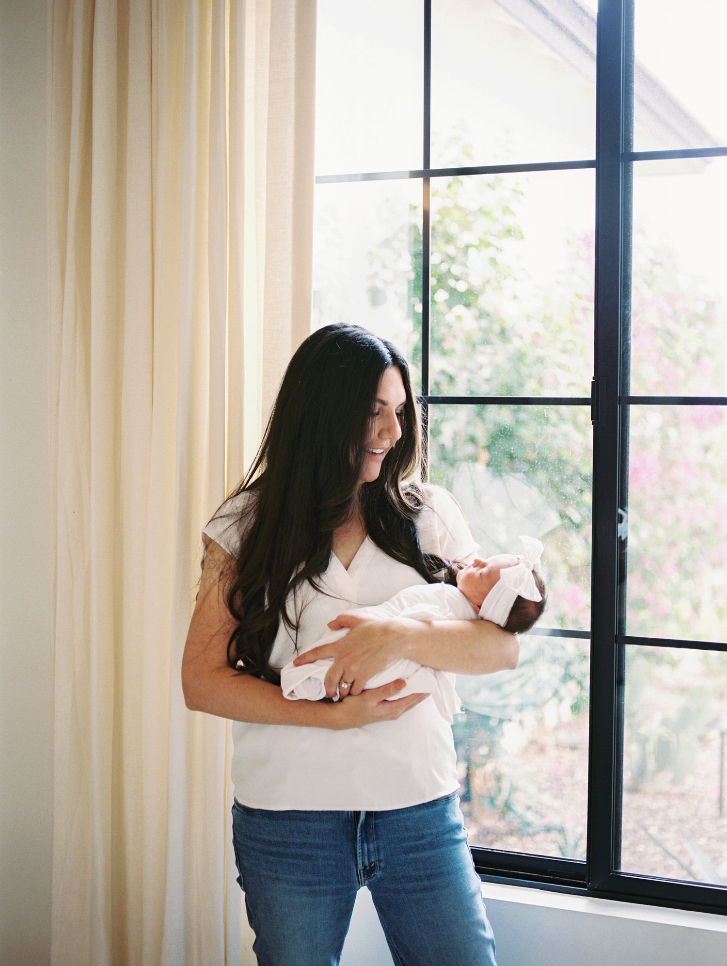 Dark-haired mother in white top and jeans cradling newborn baby by large window with natural light.