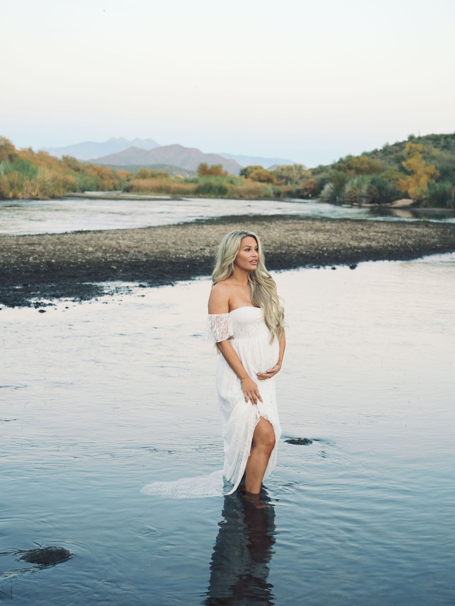 Pregnant blonde woman in white off-shoulder gown standing in shallow river with mountain backdrop.