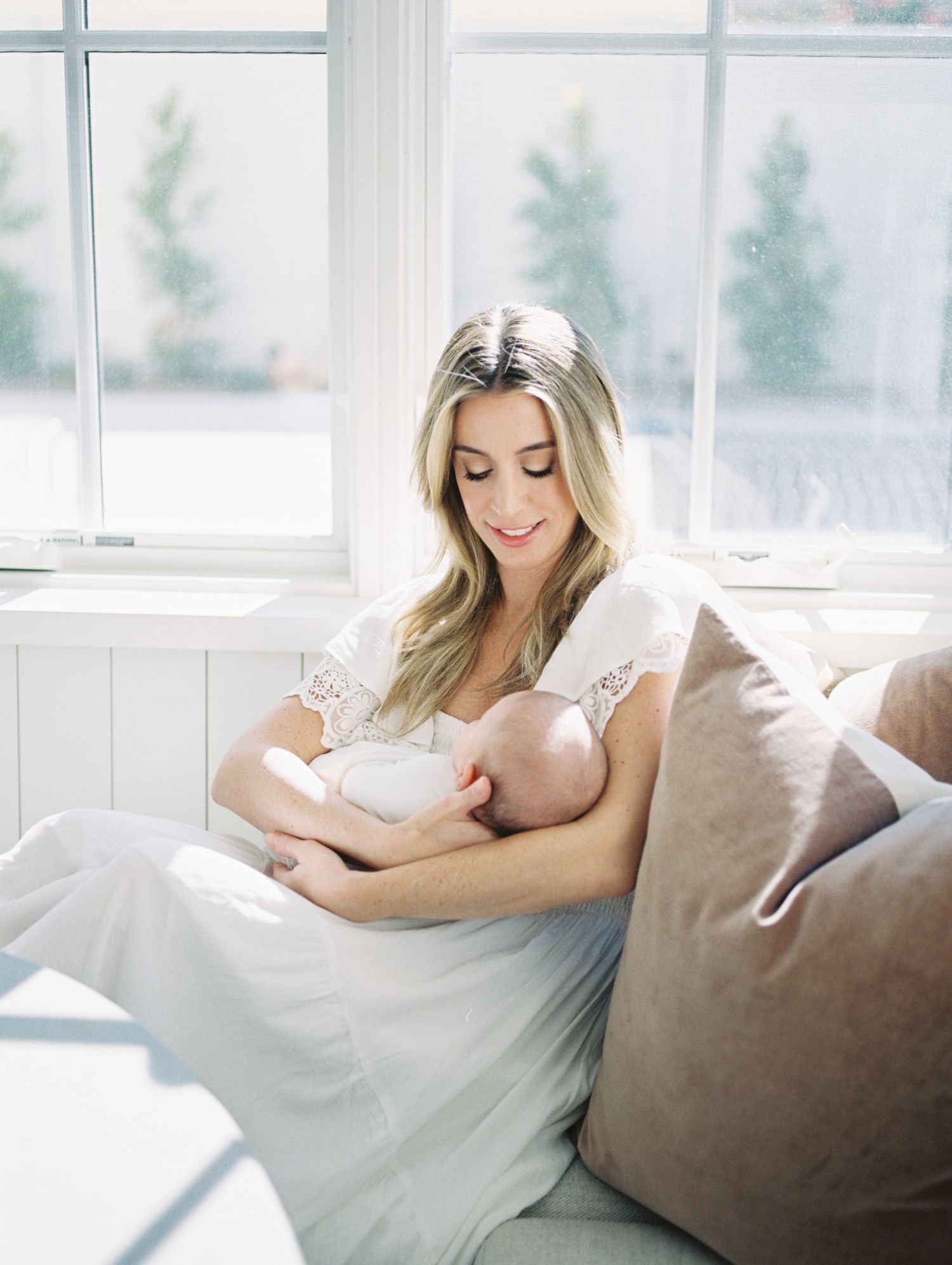 Mother in white lace dress breastfeeding newborn baby while sitting on couch near bright window.
