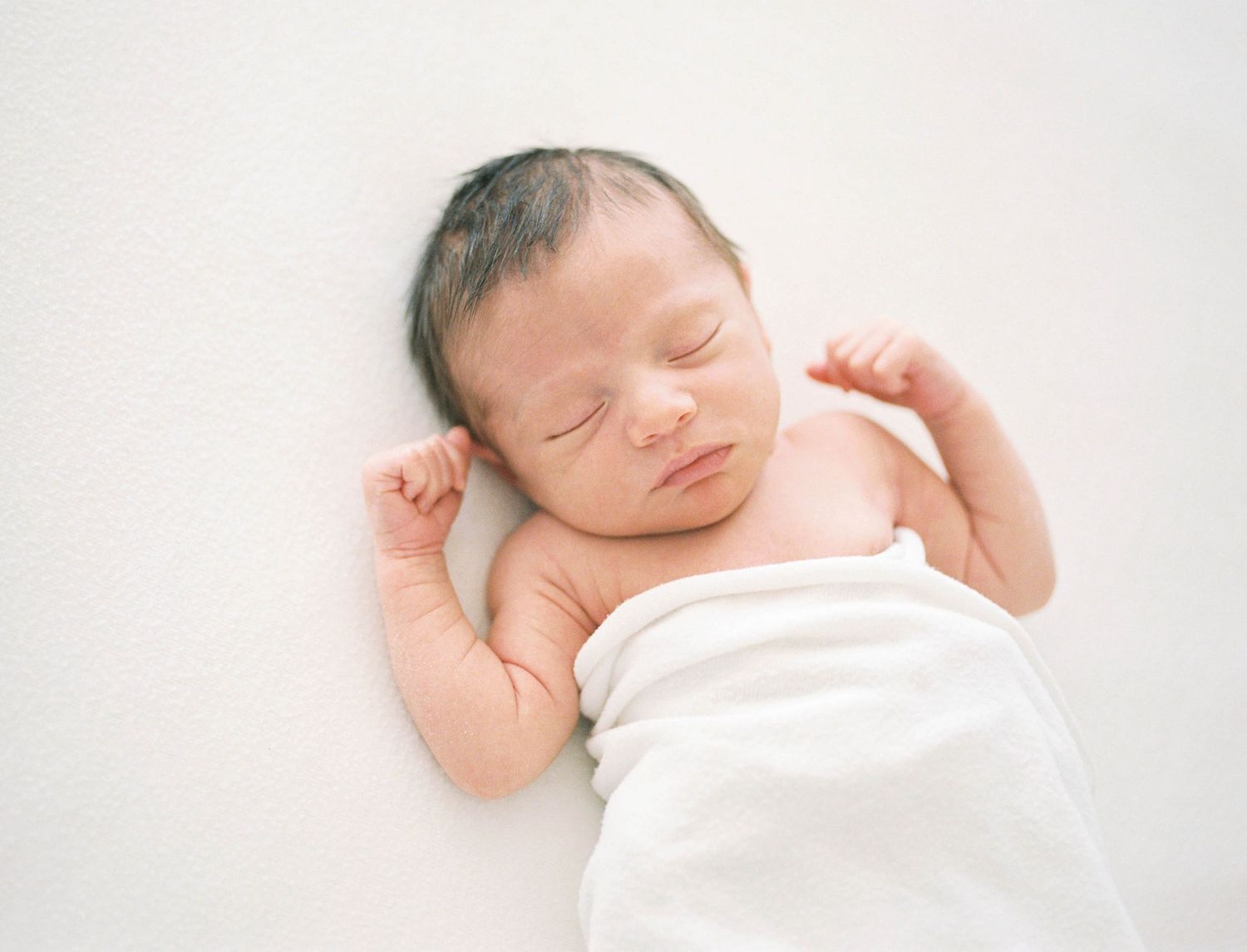 Peaceful newborn baby sleeping swaddled in white wrap on soft white background.